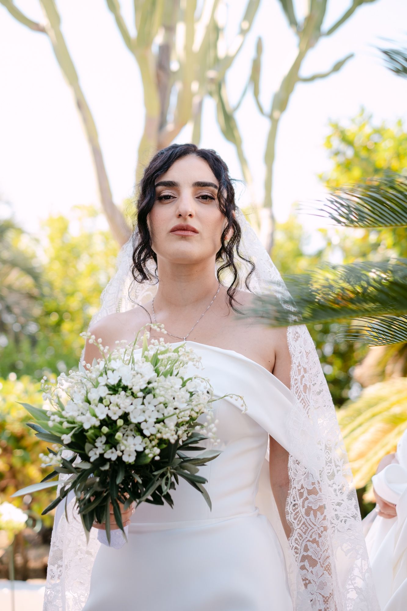 Bride in off-shoulder gown holding white bouquet outdoors with tropical foliage background.