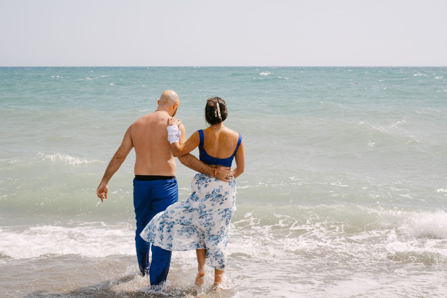 A couple walks together into the ocean waves on a sunny beach day while holding hands.