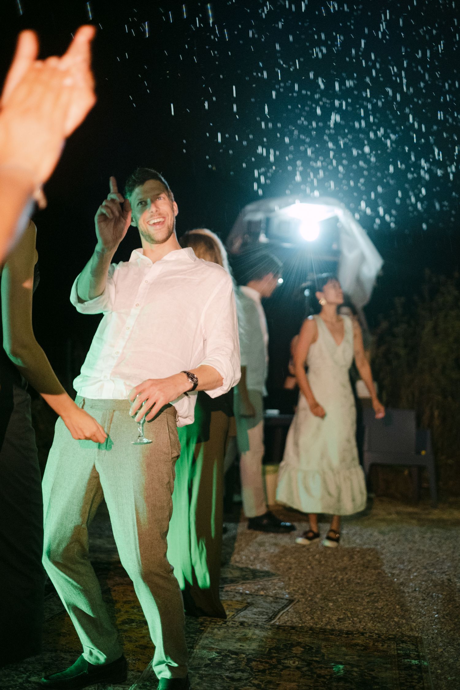 People dance under falling rain and green lighting at an outdoor nighttime celebration.