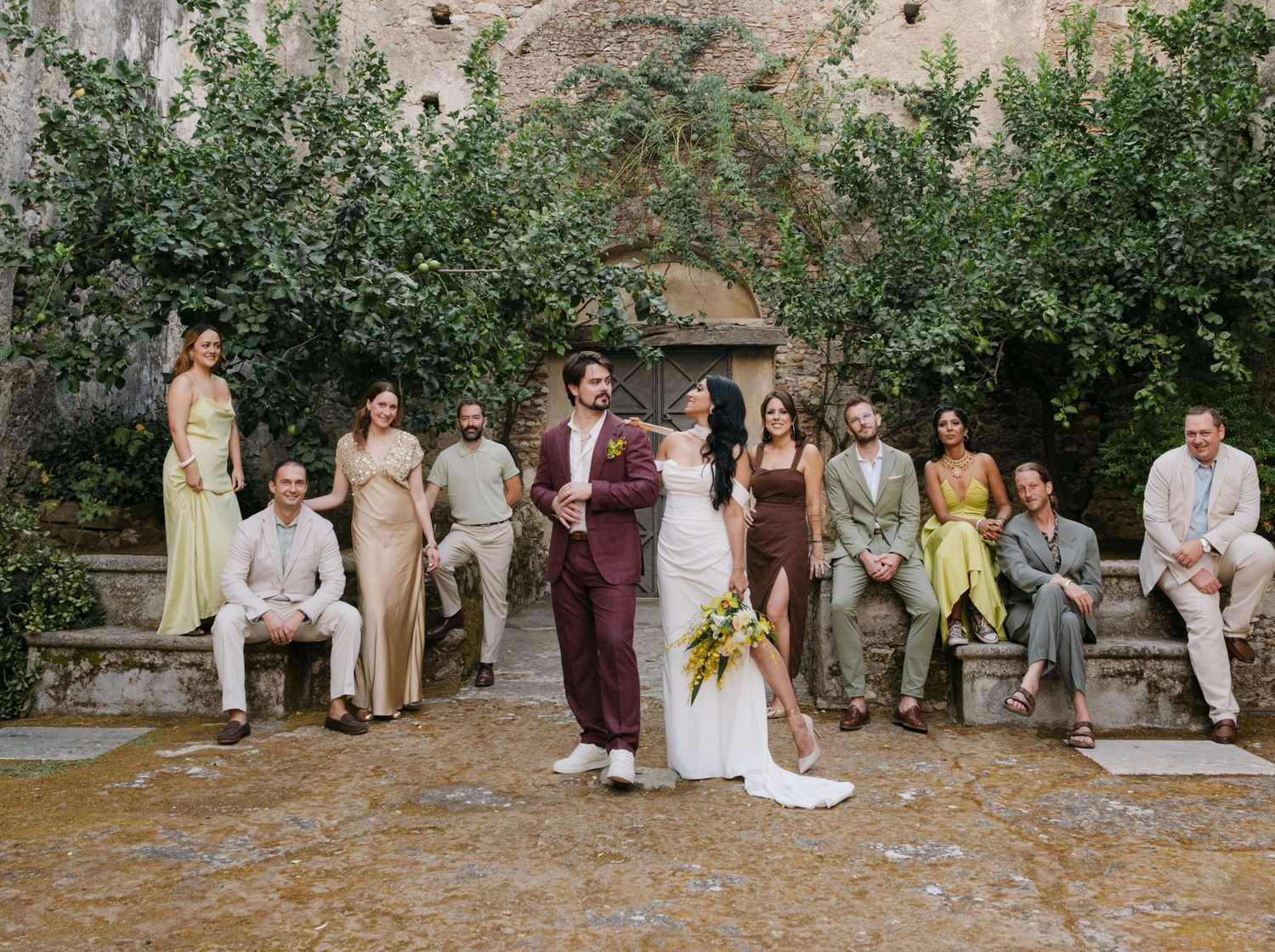 Wedding party seated on stone steps in a Mediterranean garden setting with olive trees and natural light.