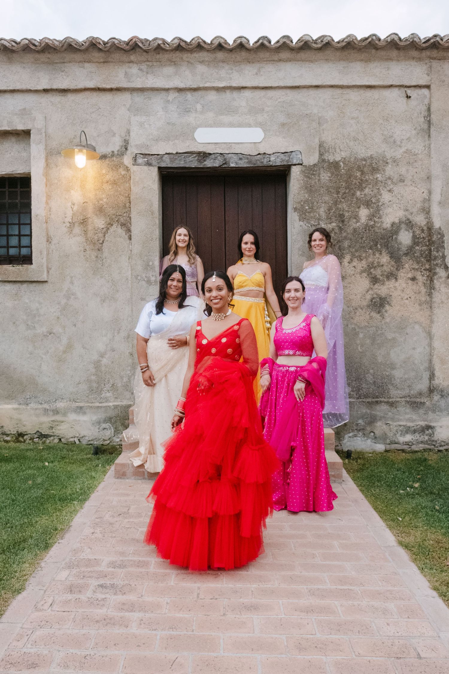 A group in vibrant red and pink formal dresses poses together on a stone patio against a weathered wall.