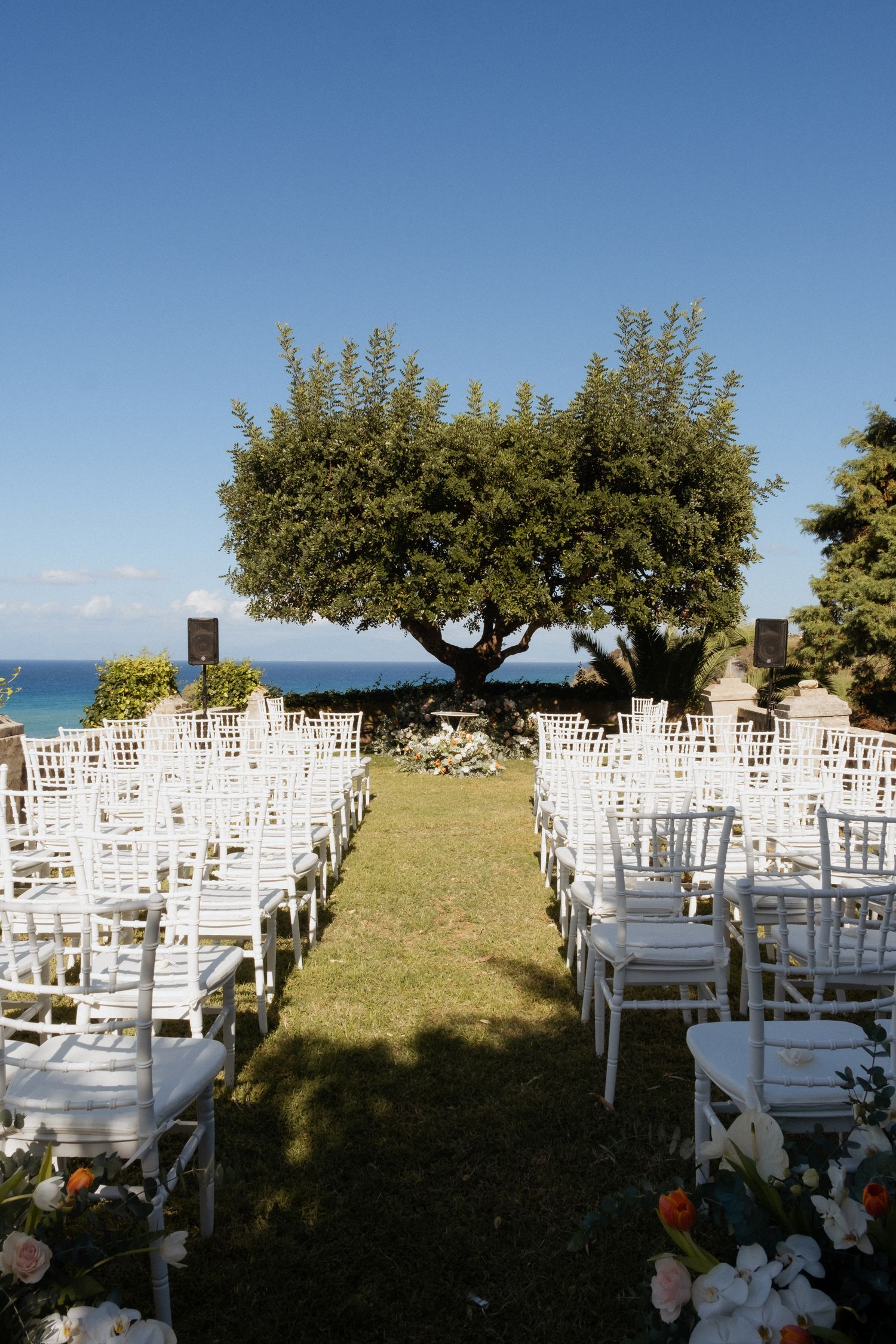 White wooden chairs arranged for an outdoor wedding ceremony overlooking the ocean under shady trees.