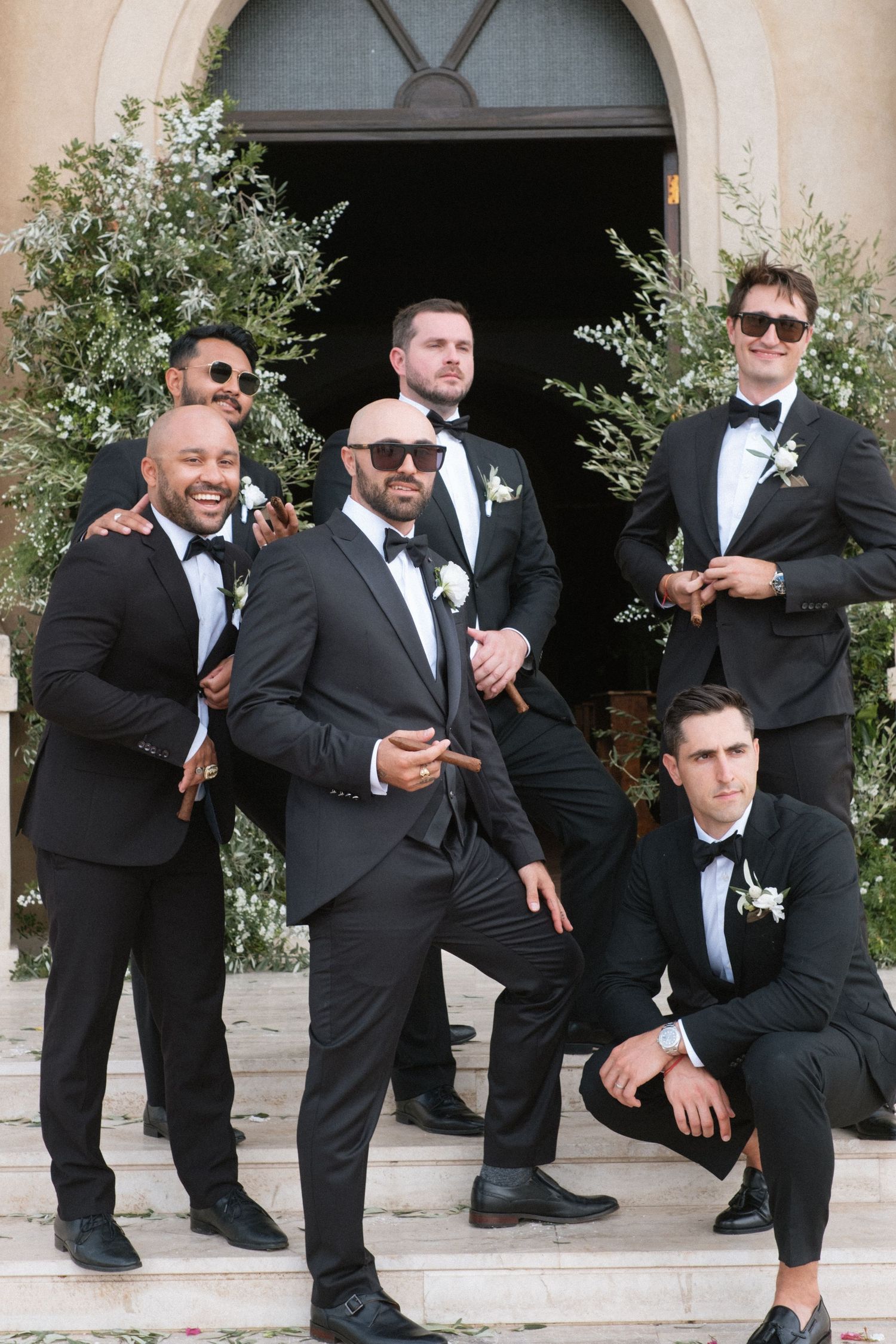 Groomsmen in black tuxedos striking fun poses on stone steps.