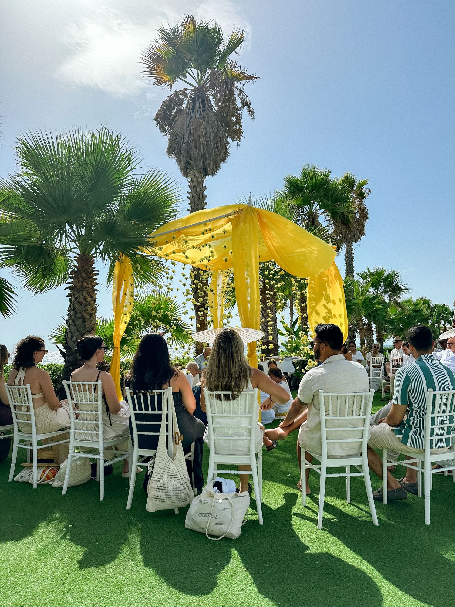 Wedding guests seated in white chairs under yellow canopies on a grassy lawn with palm trees in the background.
