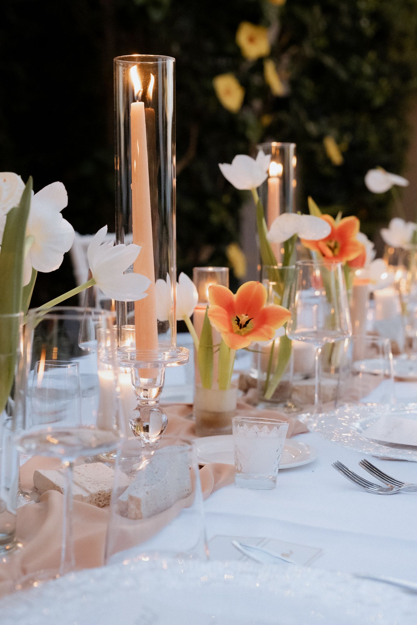Elegant wedding table setting with white tulips, orange poppies, and glass candle holders on a white tablecloth.