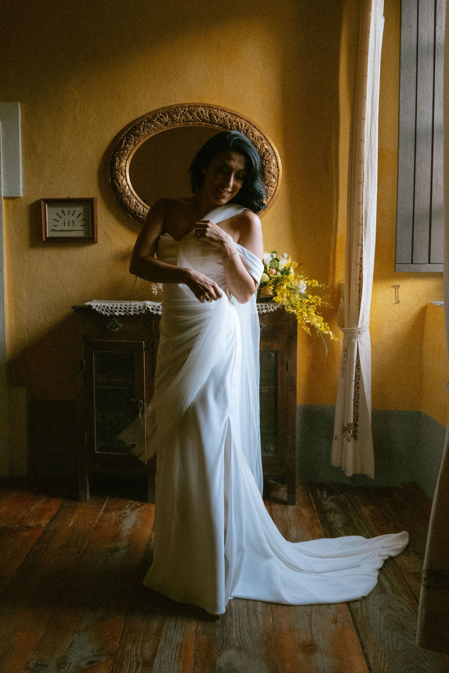 A bride in a flowing white gown poses elegantly against a golden wall with an ornate mirror.