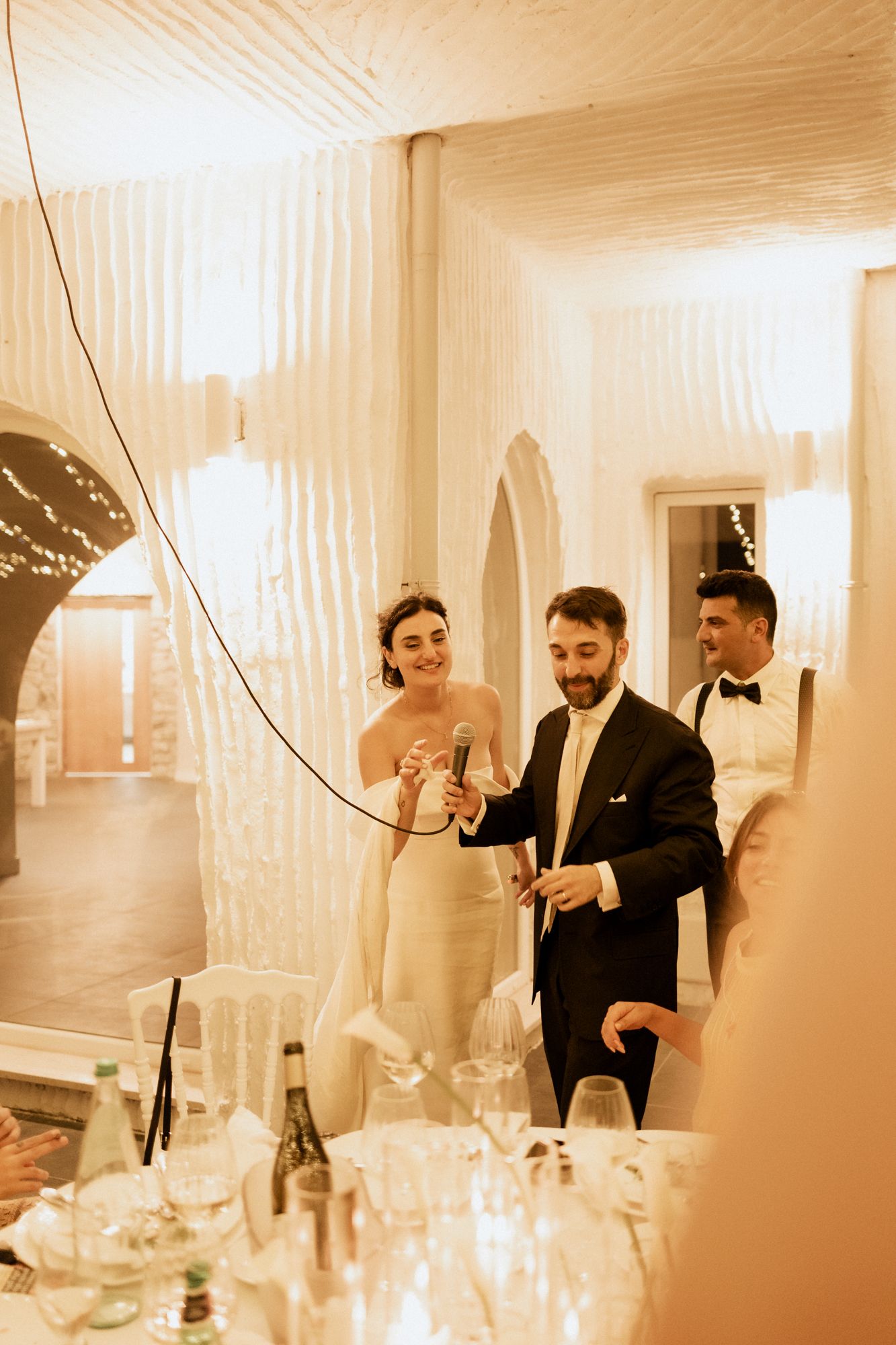 Wedding reception scene with guests in formal attire mingling in an elegantly lit white interior space.