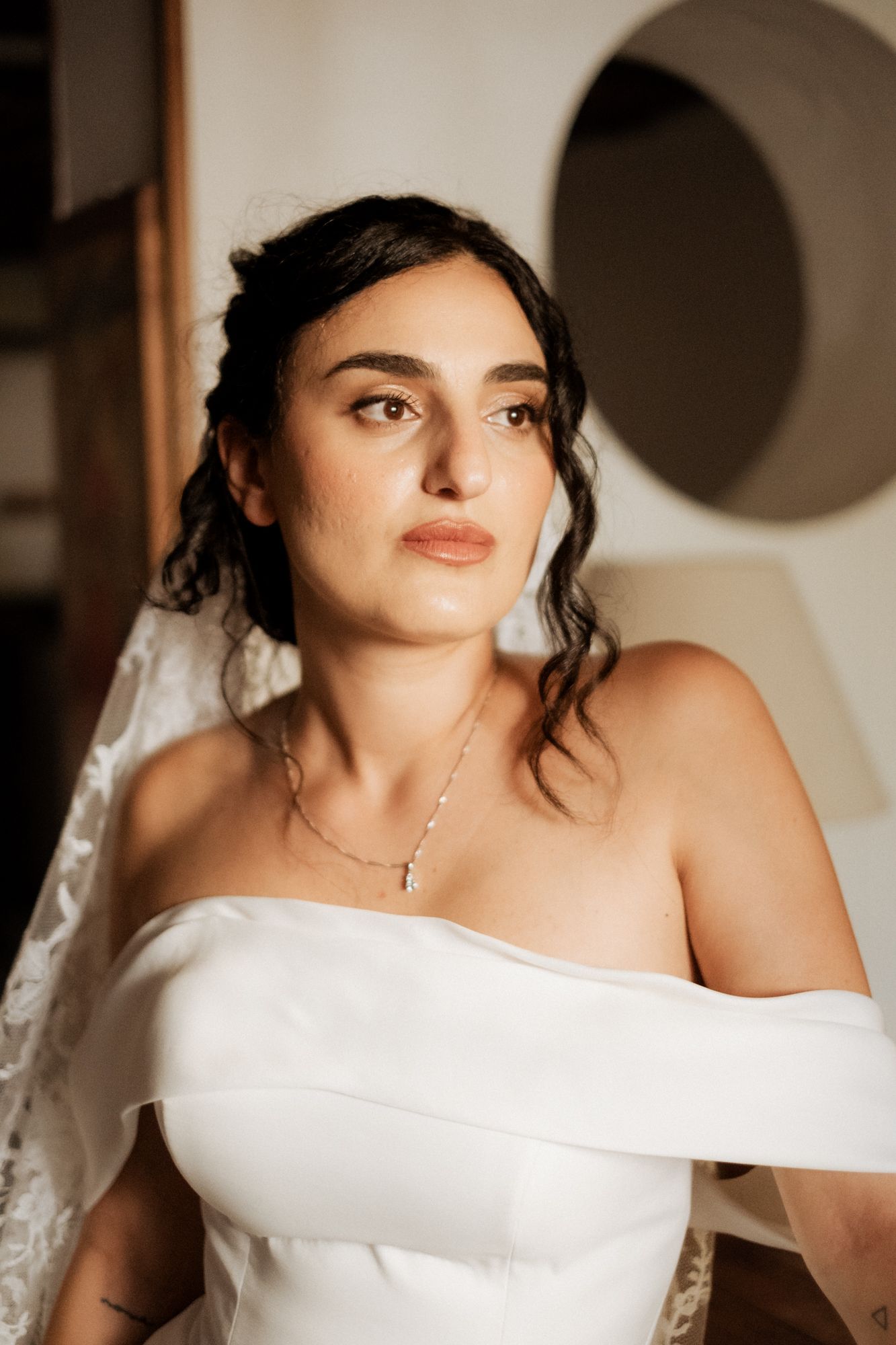 A bride in an off-shoulder wedding gown poses for intimate portraits indoors.