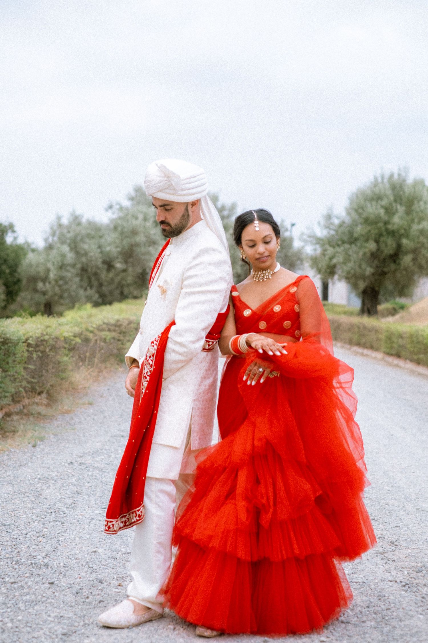 A couple in traditional attire poses on a gravel path surrounded by olive trees, with the person wearing a red gown.