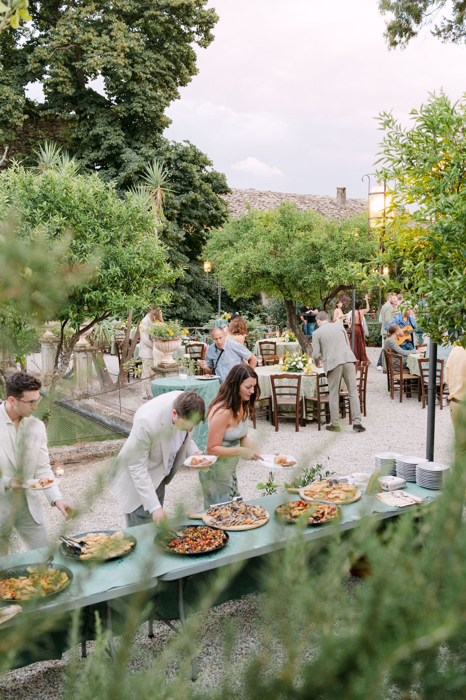 Long buffet table with appetizers and food set up outdoors under green trees.