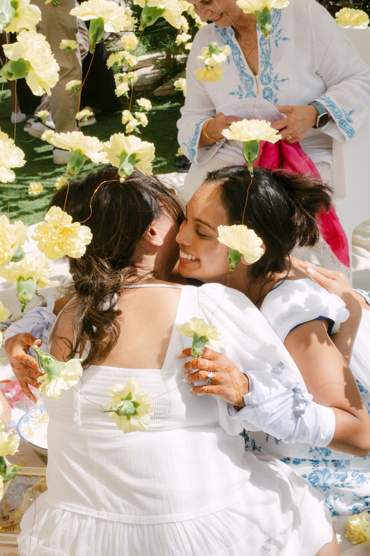 Two people in white dresses embrace joyfully surrounded by delicate cream-colored flowers at a celebration.