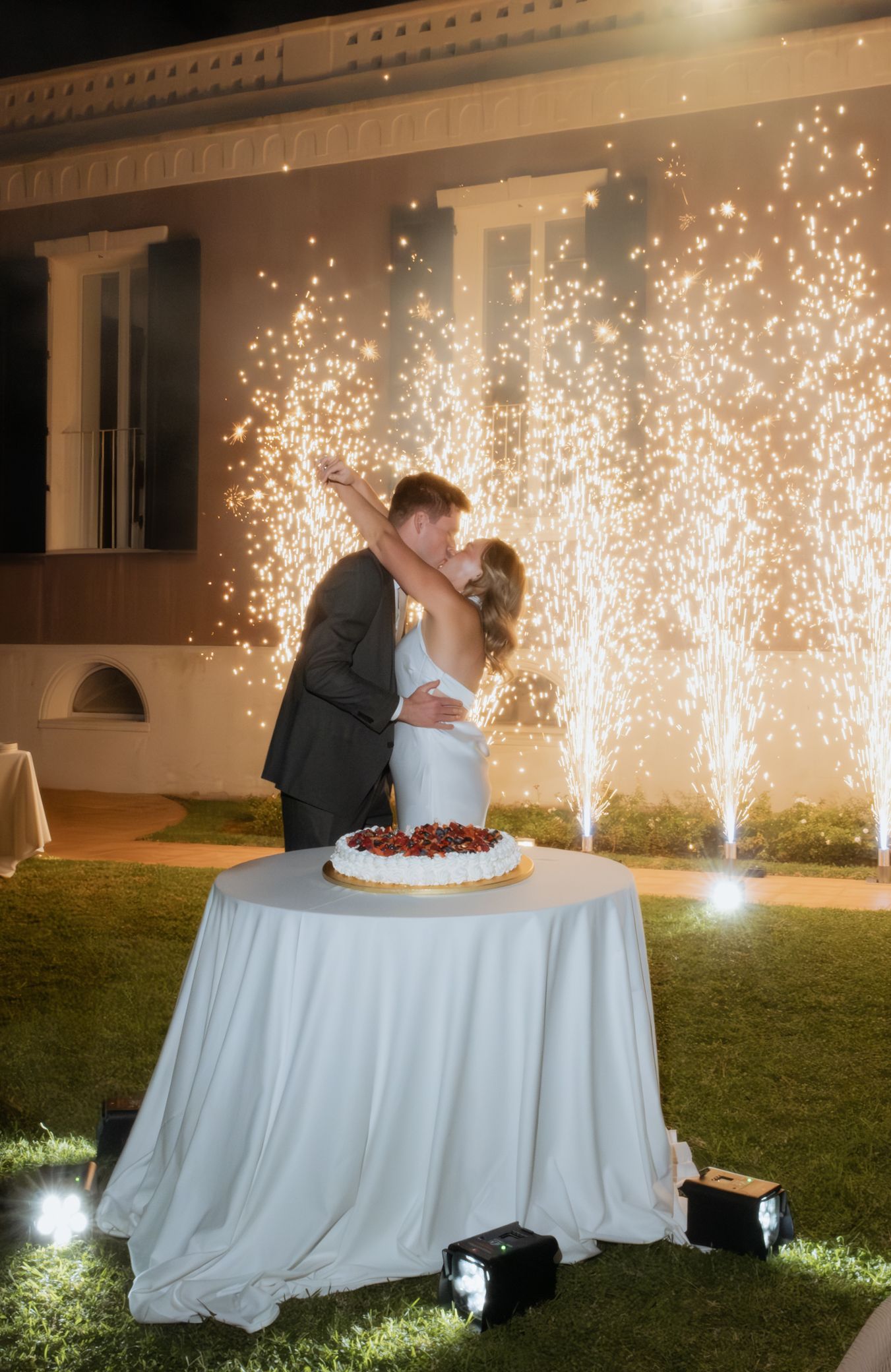 Wedding couple cuts their cake outdoors with dramatic sparkler fountains illuminating the background.