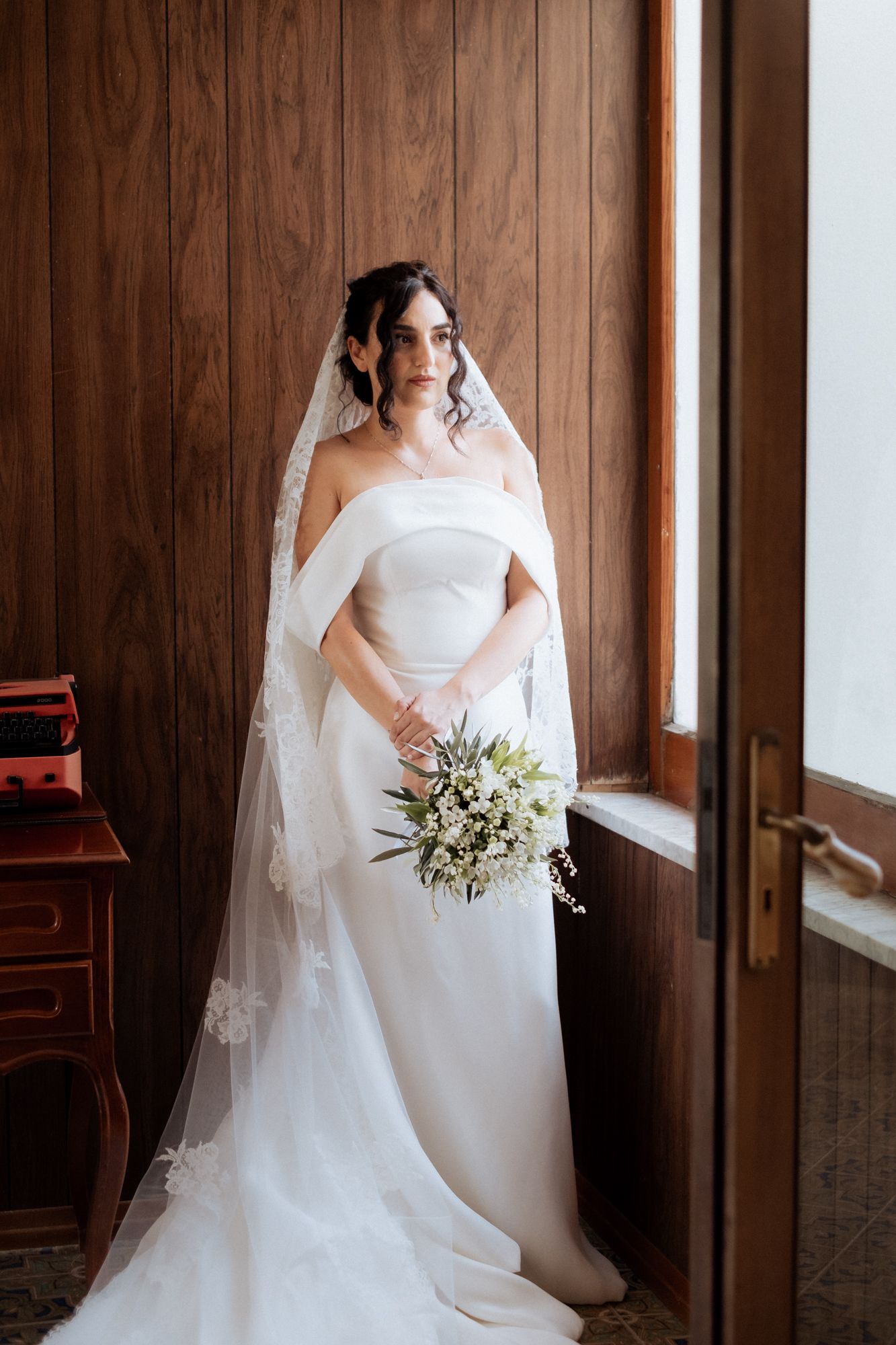 A bride in a classic white wedding gown holds a delicate bouquet in a wood-paneled room.