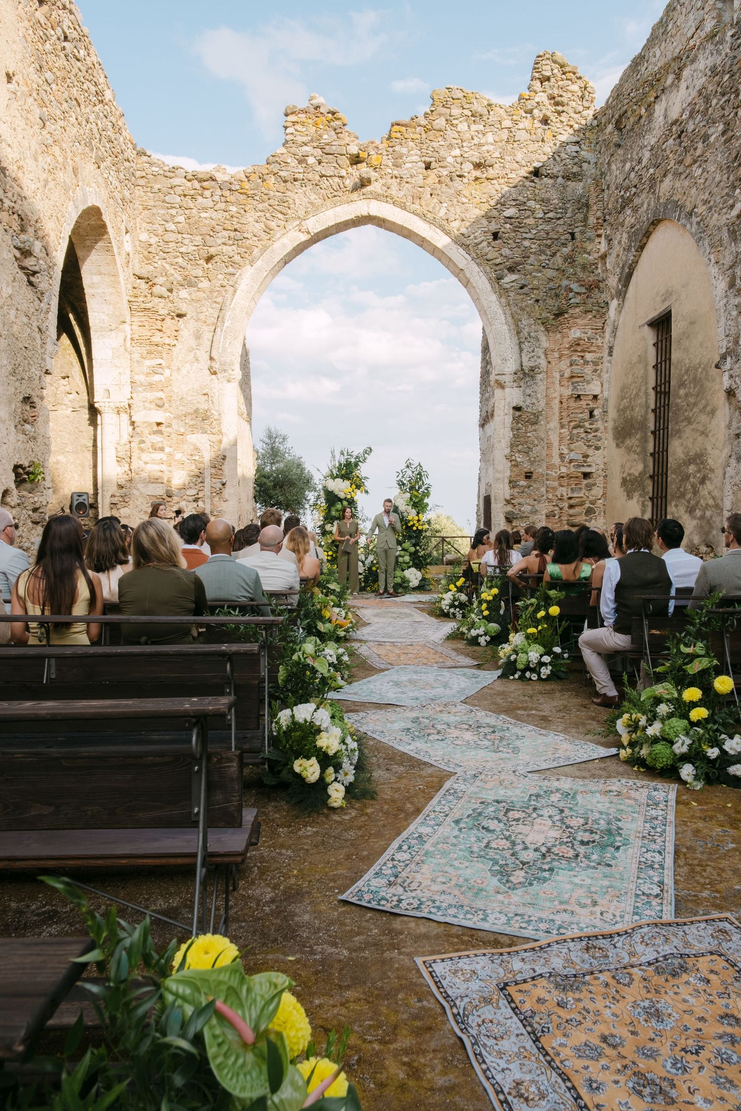 Ancient stone ruins decorated with floral arrangements and oriental rugs create an enchanting outdoor wedding ceremony space.