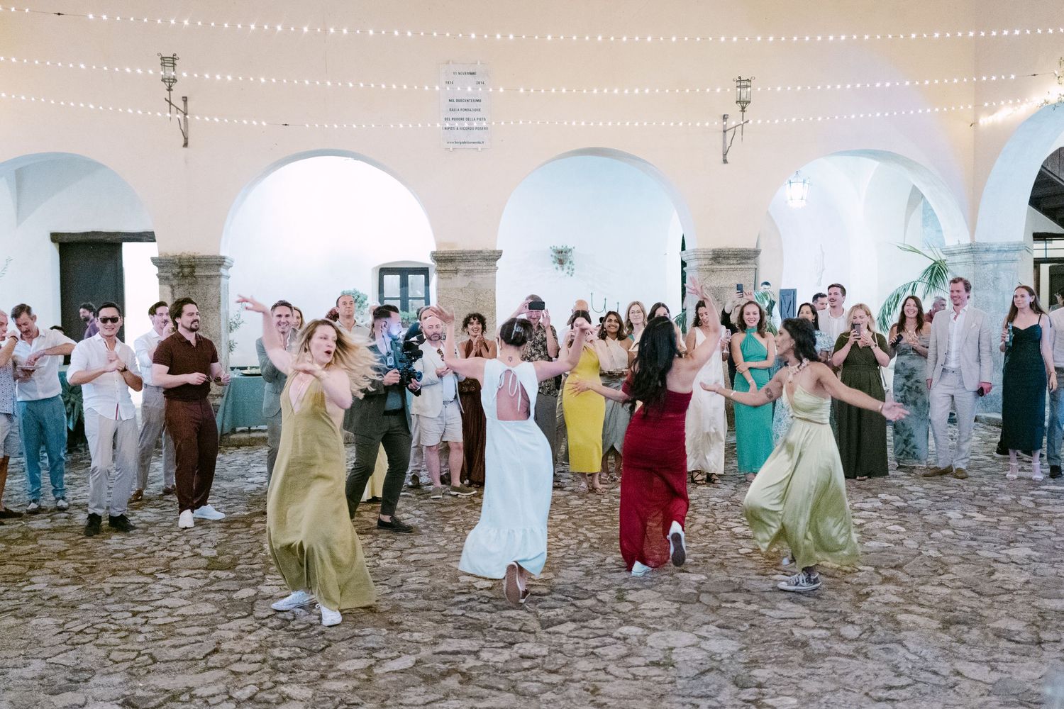 Wedding guests dancing in a line formation against white walls at an outdoor celebration.