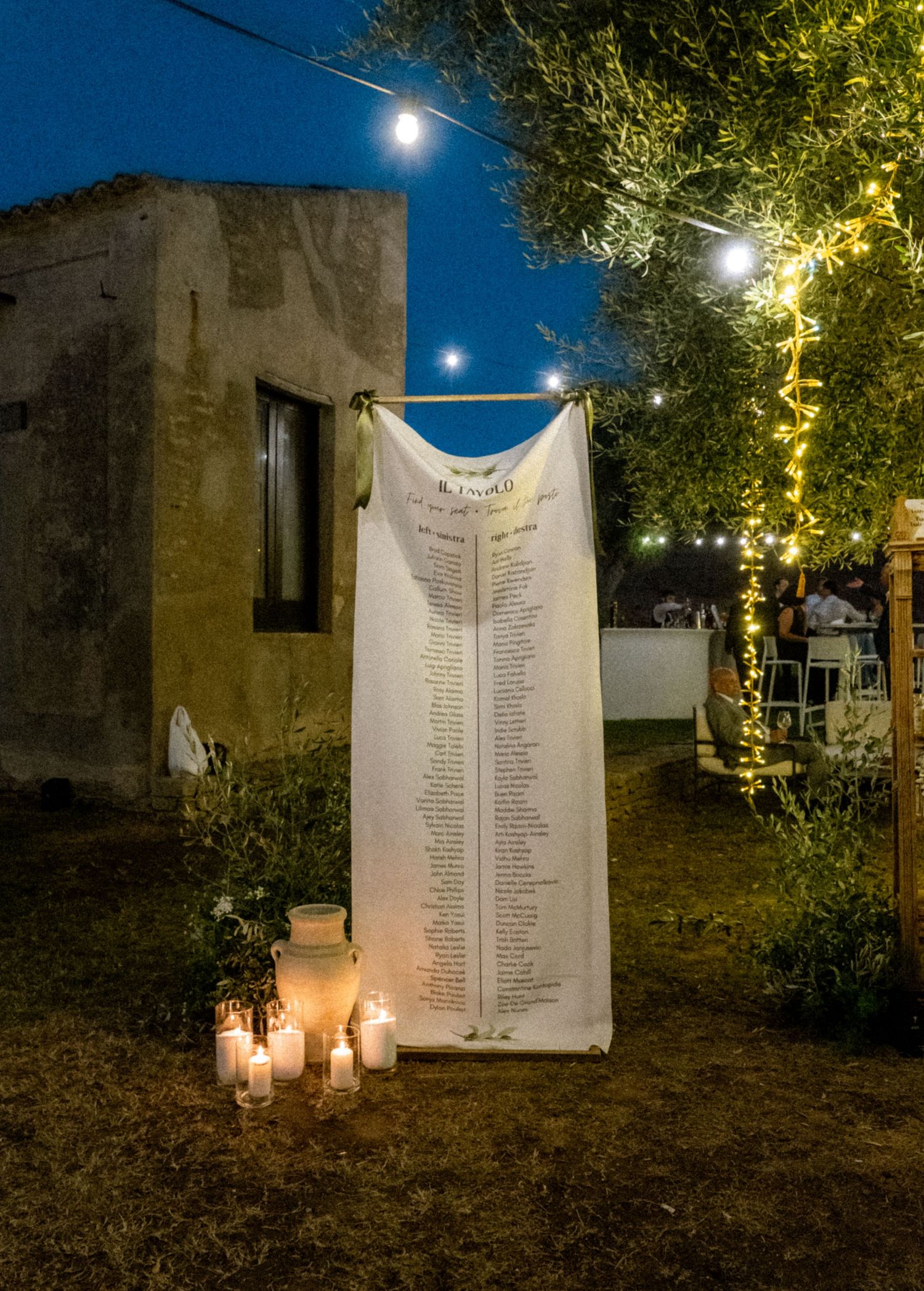 Wedding seating chart display illuminated by candlelight and string lights in an outdoor garden setting.