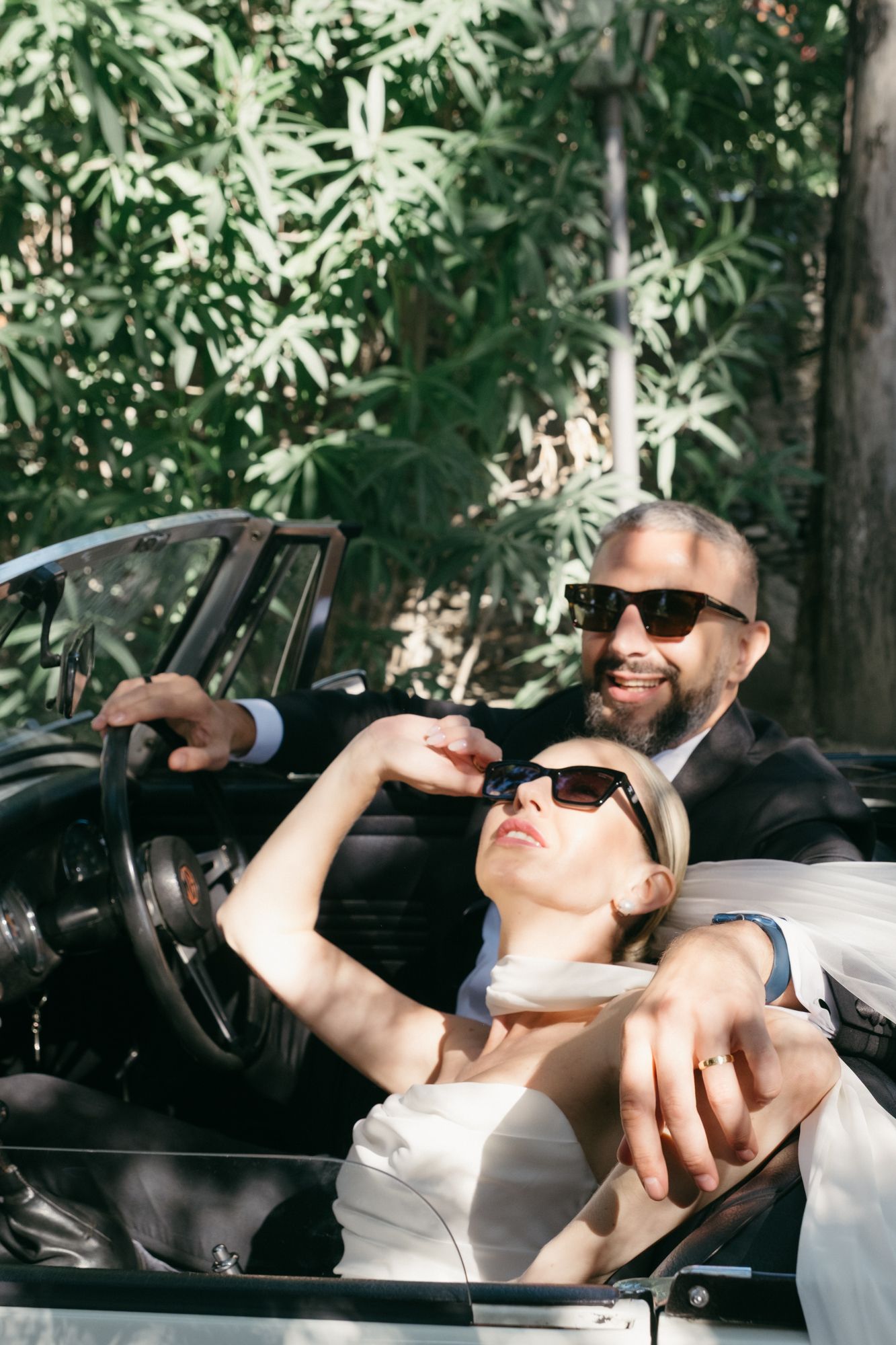People enjoying drinks in a convertible car surrounded by lush greenery on a sunny day.