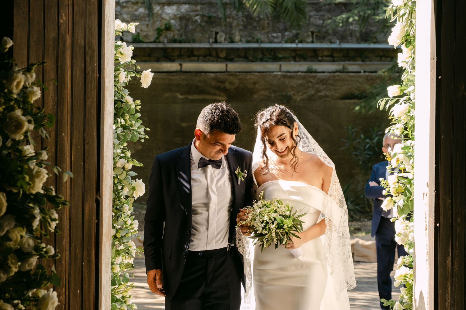 A wedding ceremony takes place under an archway decorated with greenery and flowers.