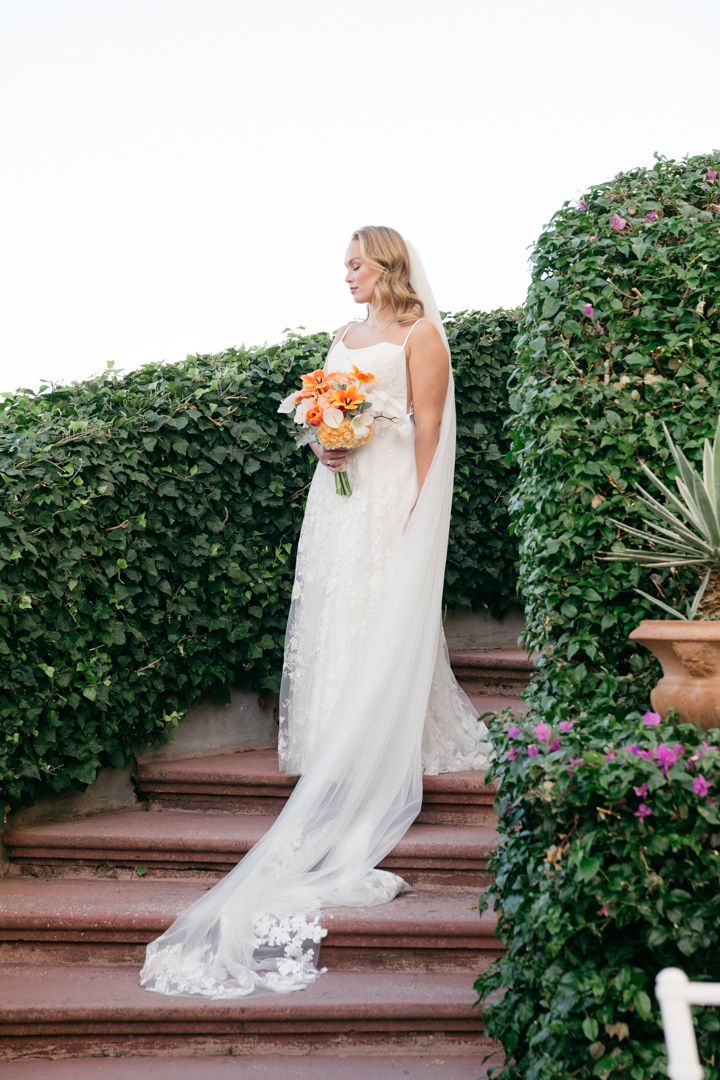 A flowing white wedding dress with lace train cascades down stone steps surrounded by lush greenery.