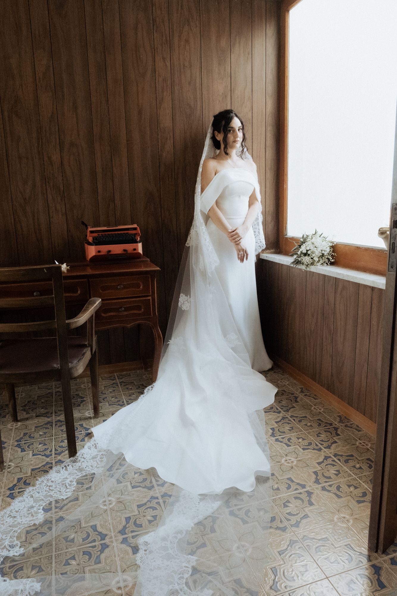 Bride in fitted white wedding dress posing by wooden wall with long lace veil.