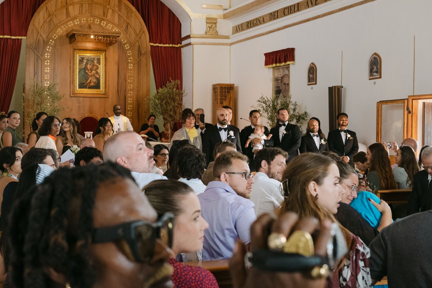 Wedding ceremony guests seated in ornate church interior with gilded altar.