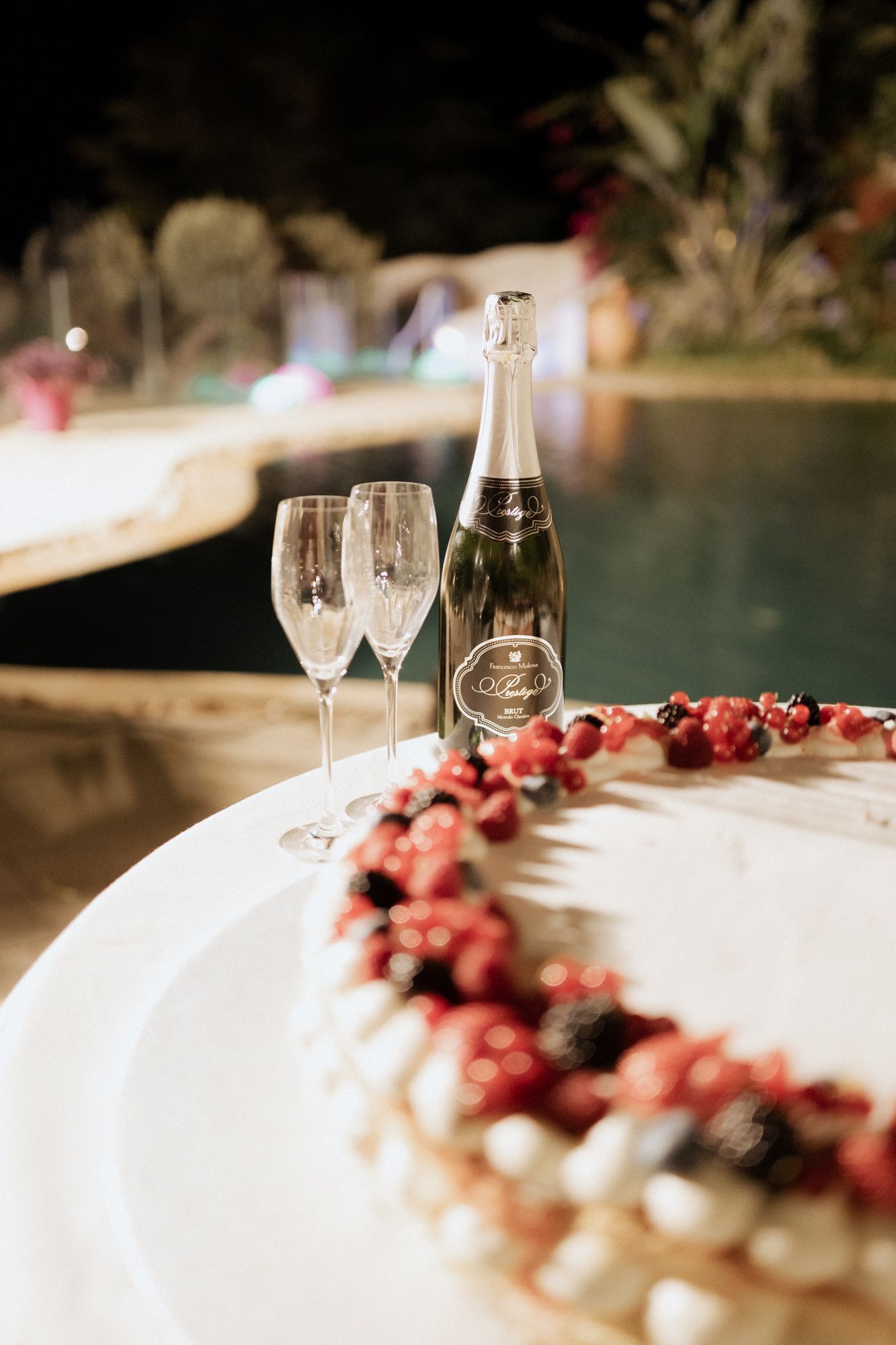 Wedding cake decorated with berries next to champagne bottle and glasses by pool at night.