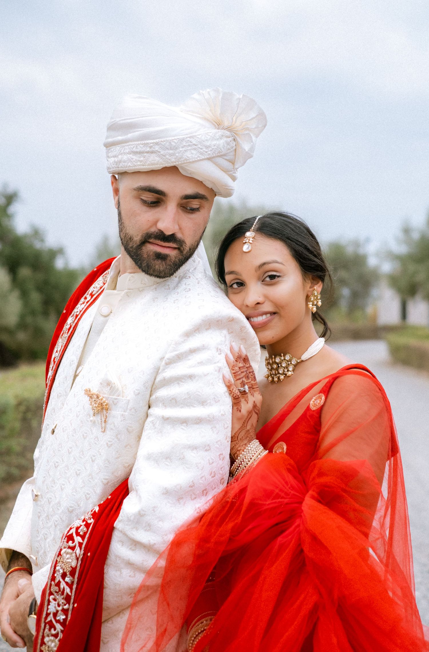 A couple in traditional Indian wedding attire pose together against an outdoor backdrop.