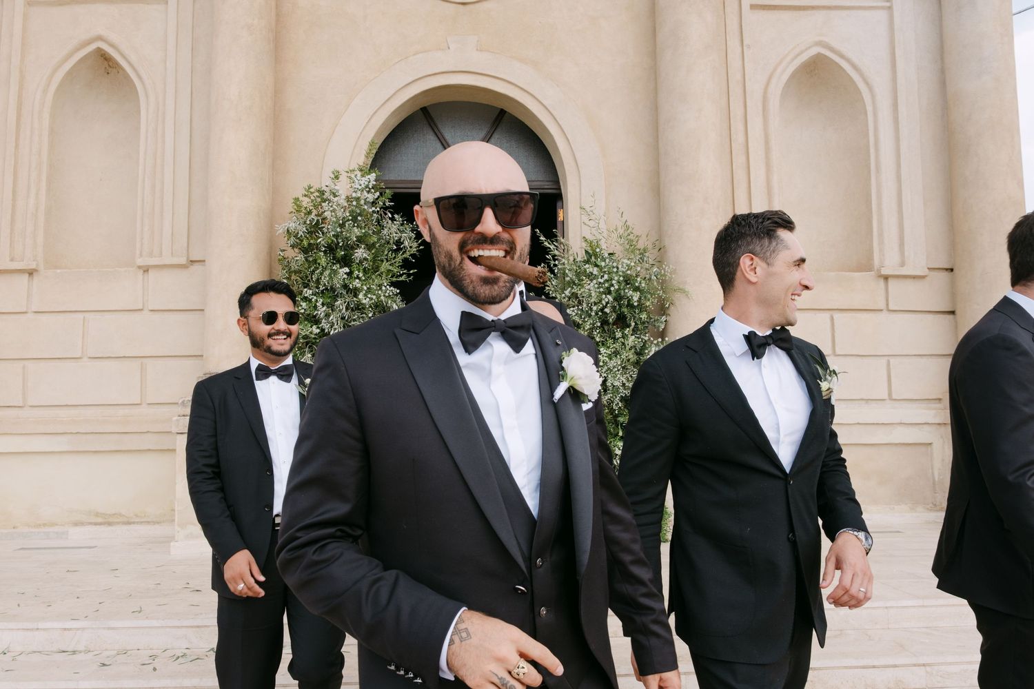 A group of groomsmen in black tuxedos and bowties laugh together during a formal wedding event.