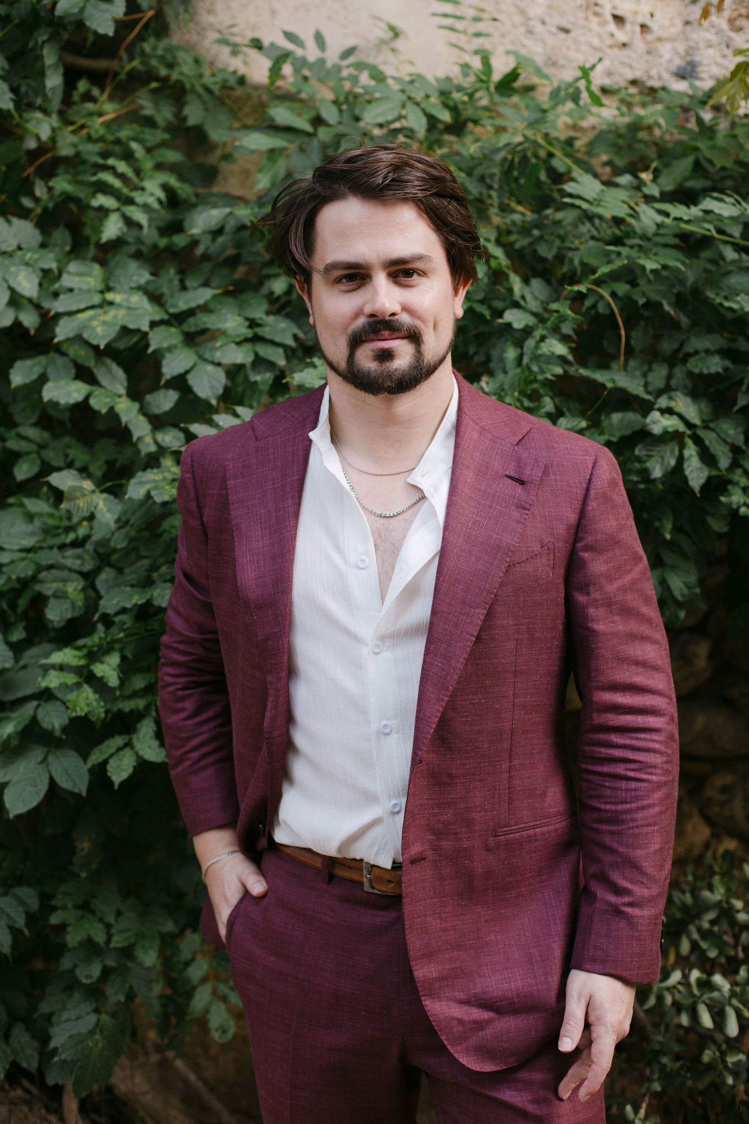 A person in a burgundy suit jacket and white shirt poses against a leafy green background.