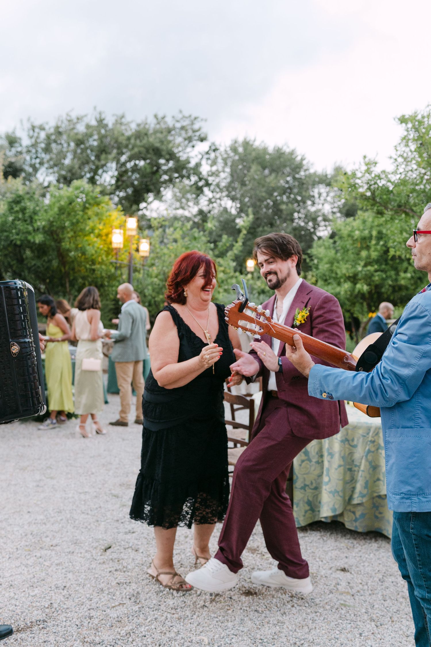 Guests mingle and chat at an outdoor event on a gravel path with trees and greenery in the background.