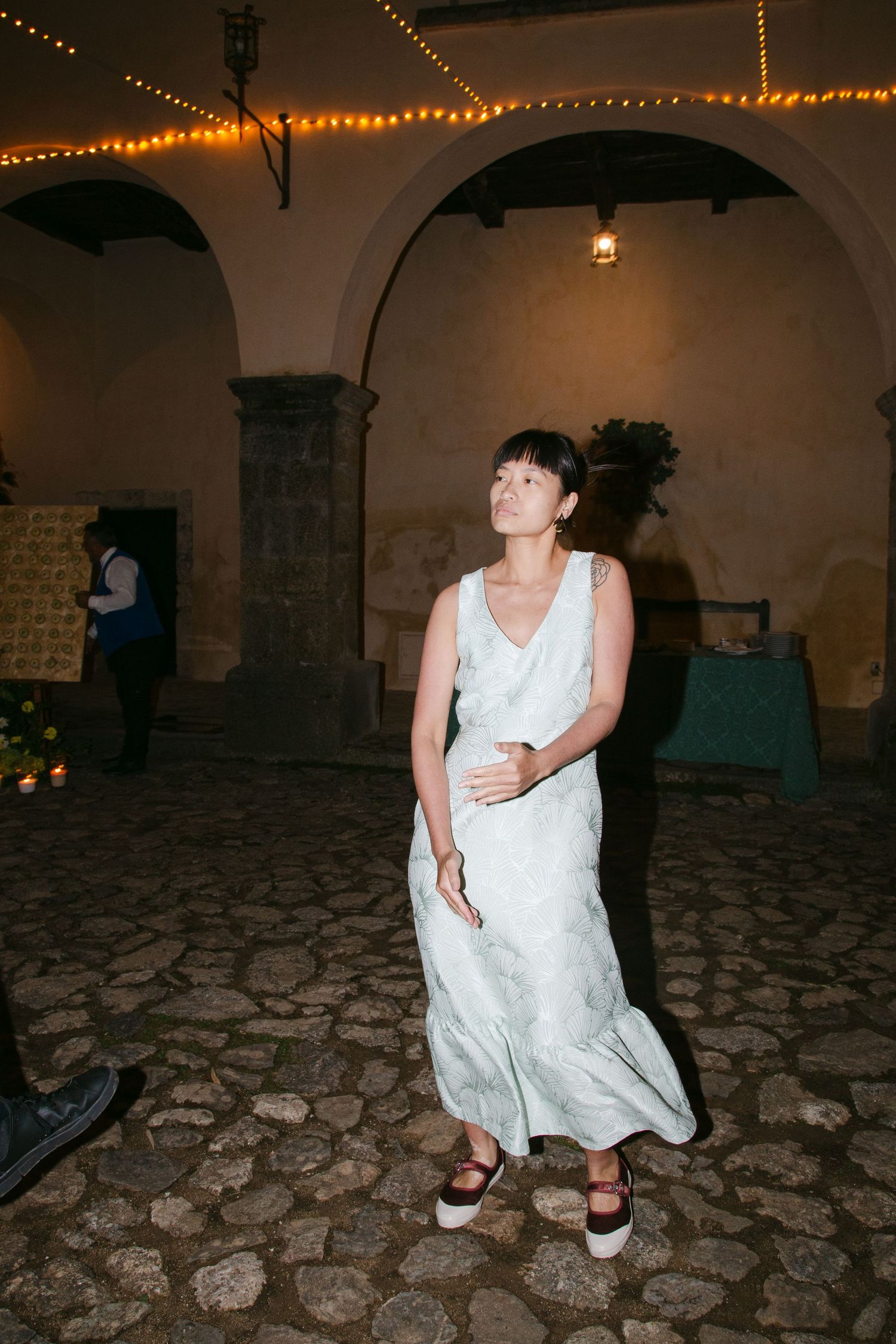 A person sits on a cobblestone floor under string lights at an evening celebration.