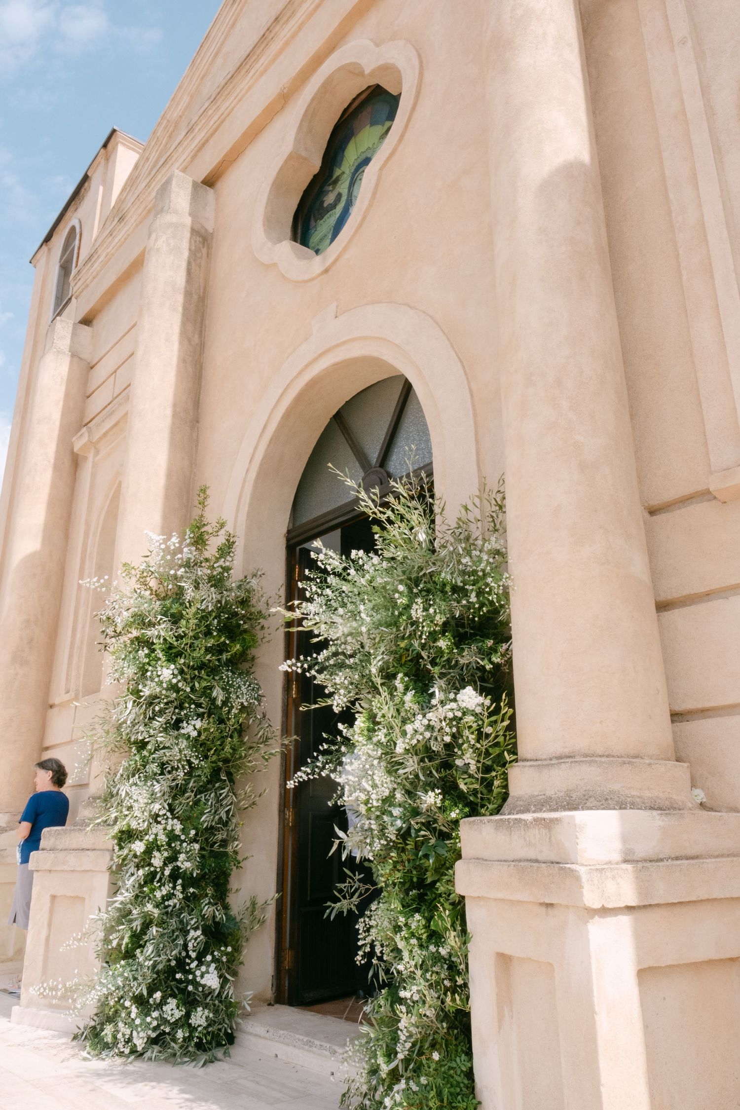 A white stone church exterior with arched windows and doorways adorned with floral arrangements.