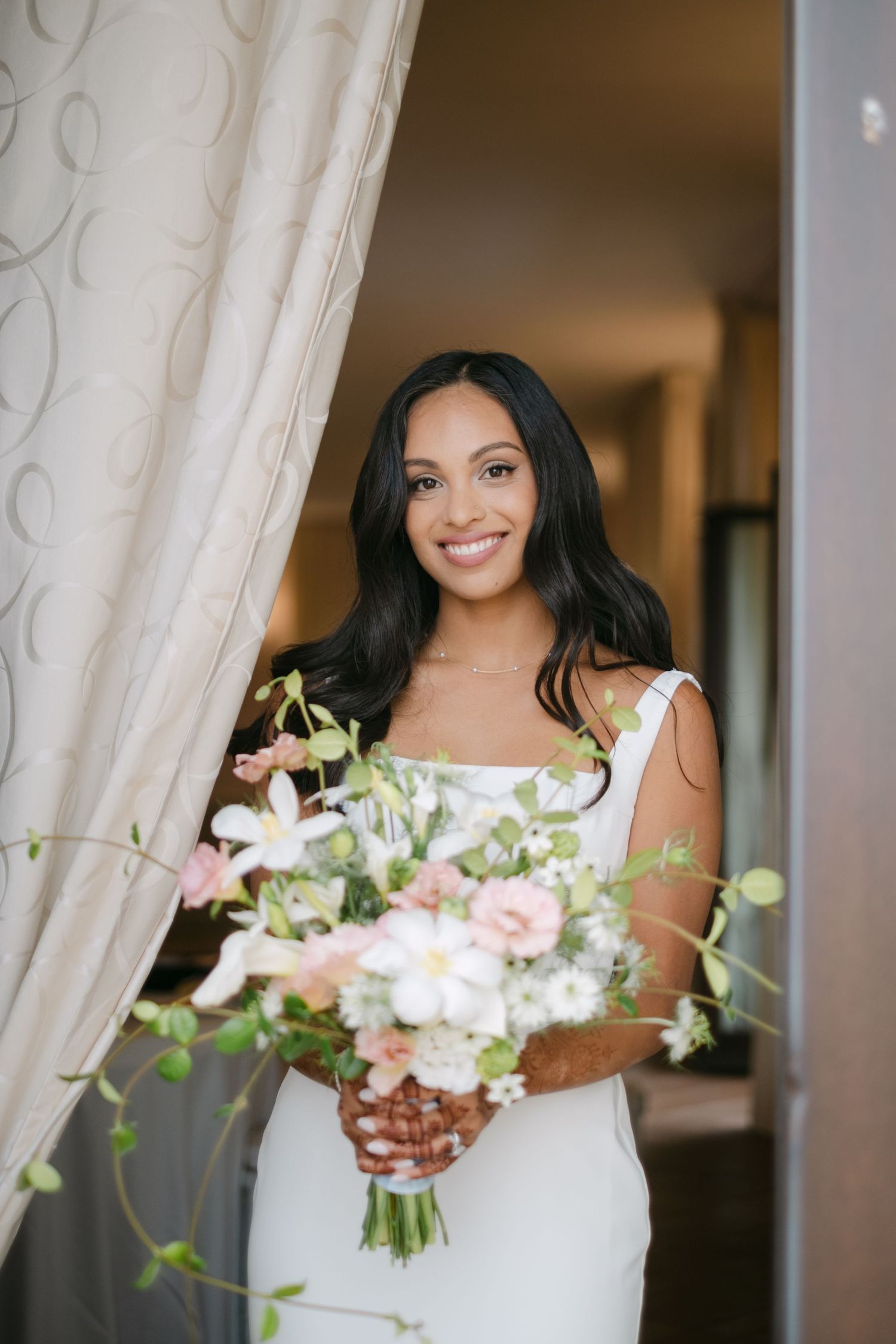 Elegant bride in white dress holding delicate floral bouquet by window with curtains.