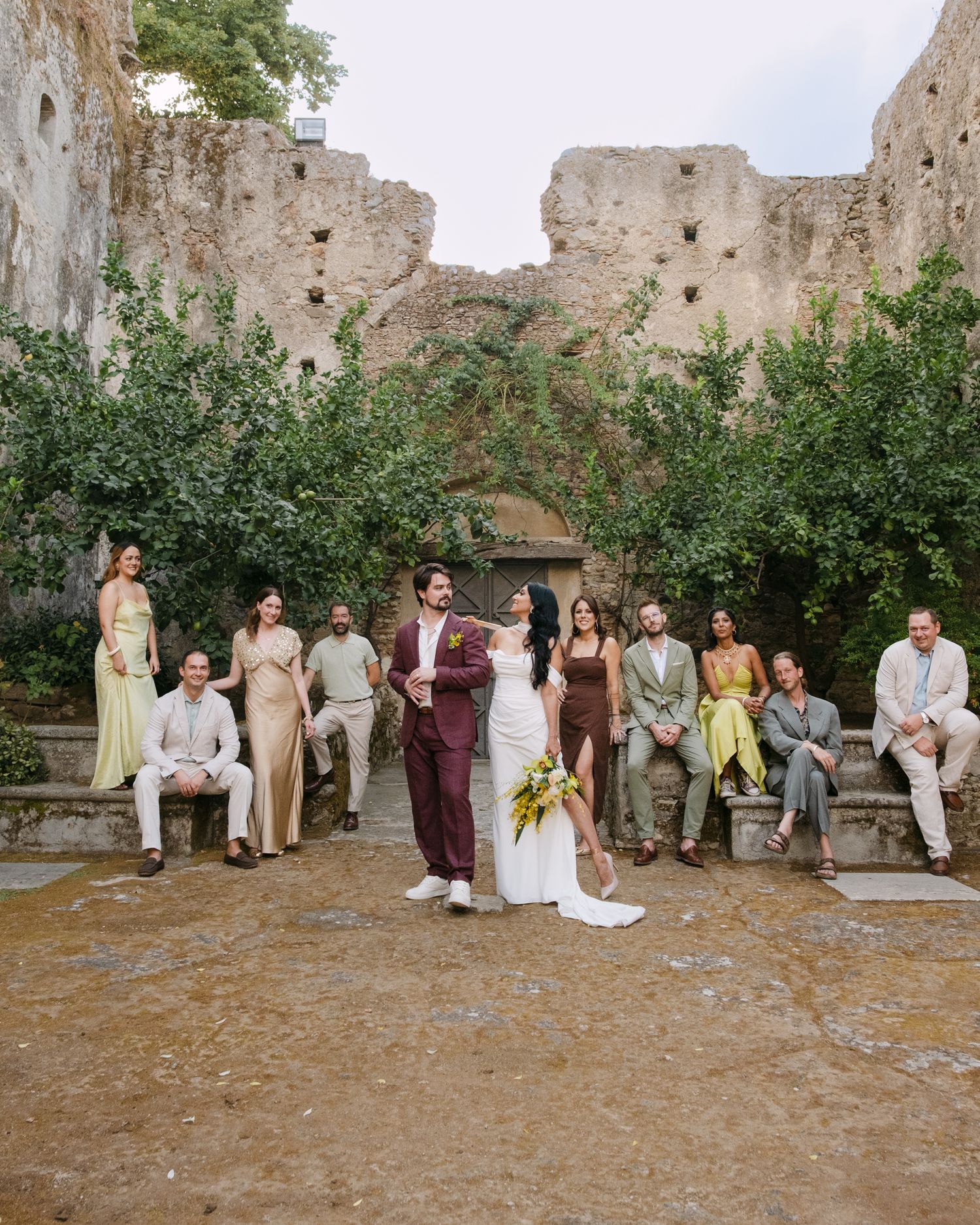 Wedding guests gather in a stone courtyard ruin surrounded by old walls and green foliage.