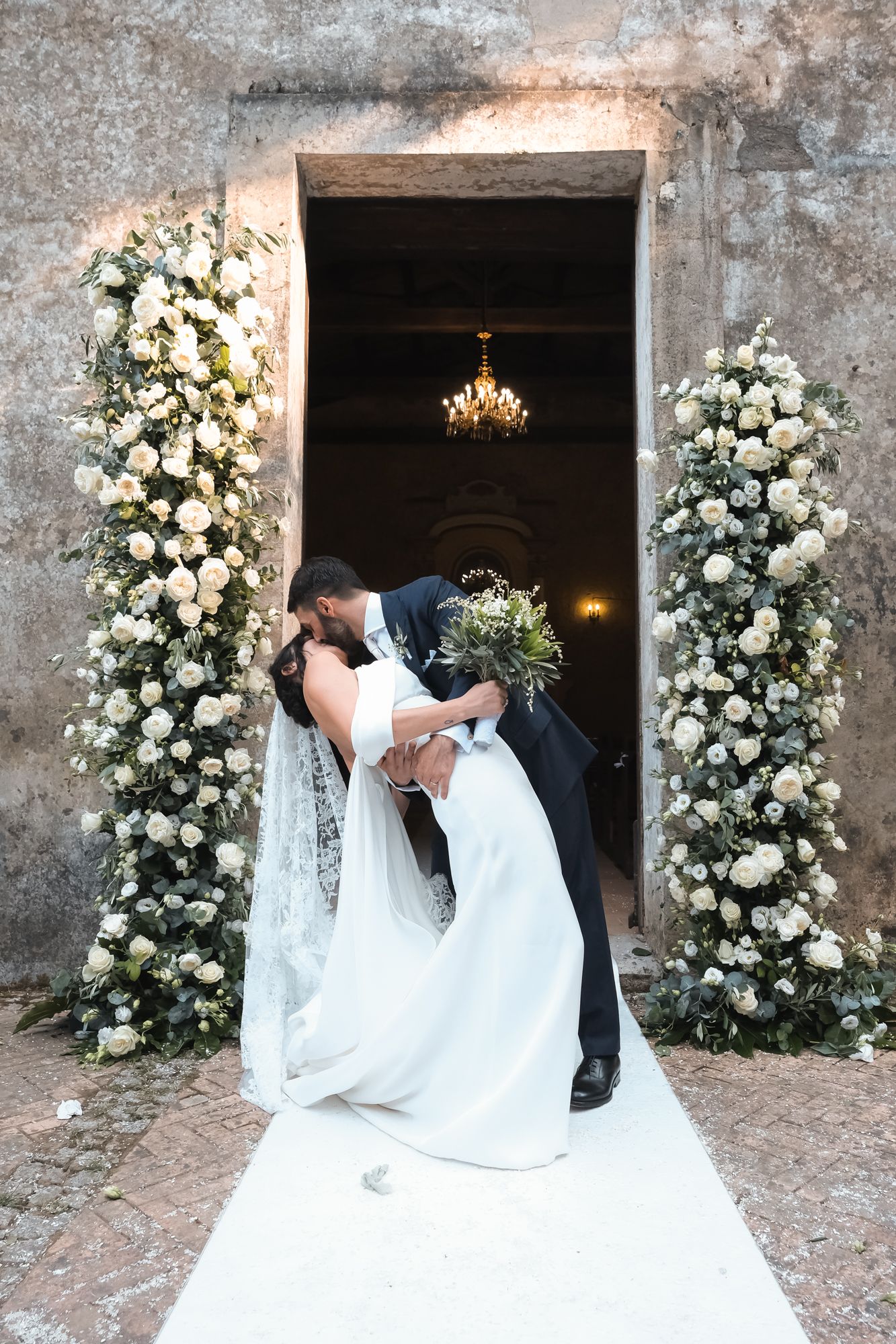 Romantic wedding couple sharing intimate kisses in front of elegant white floral arrangements against stone walls.