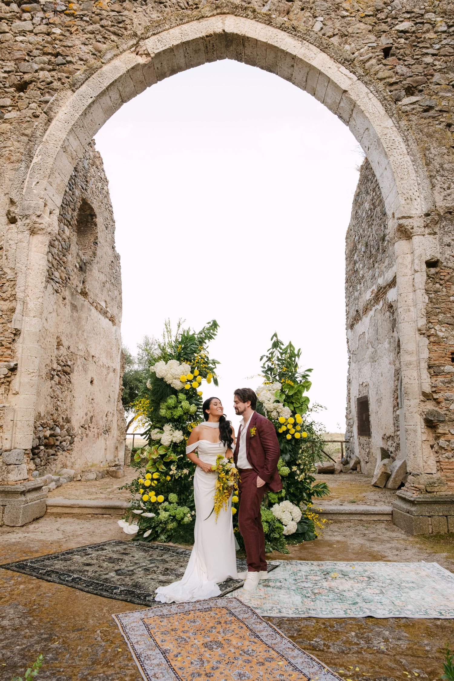 A wedding ceremony taking place under stone archways decorated with yellow and white flowers.