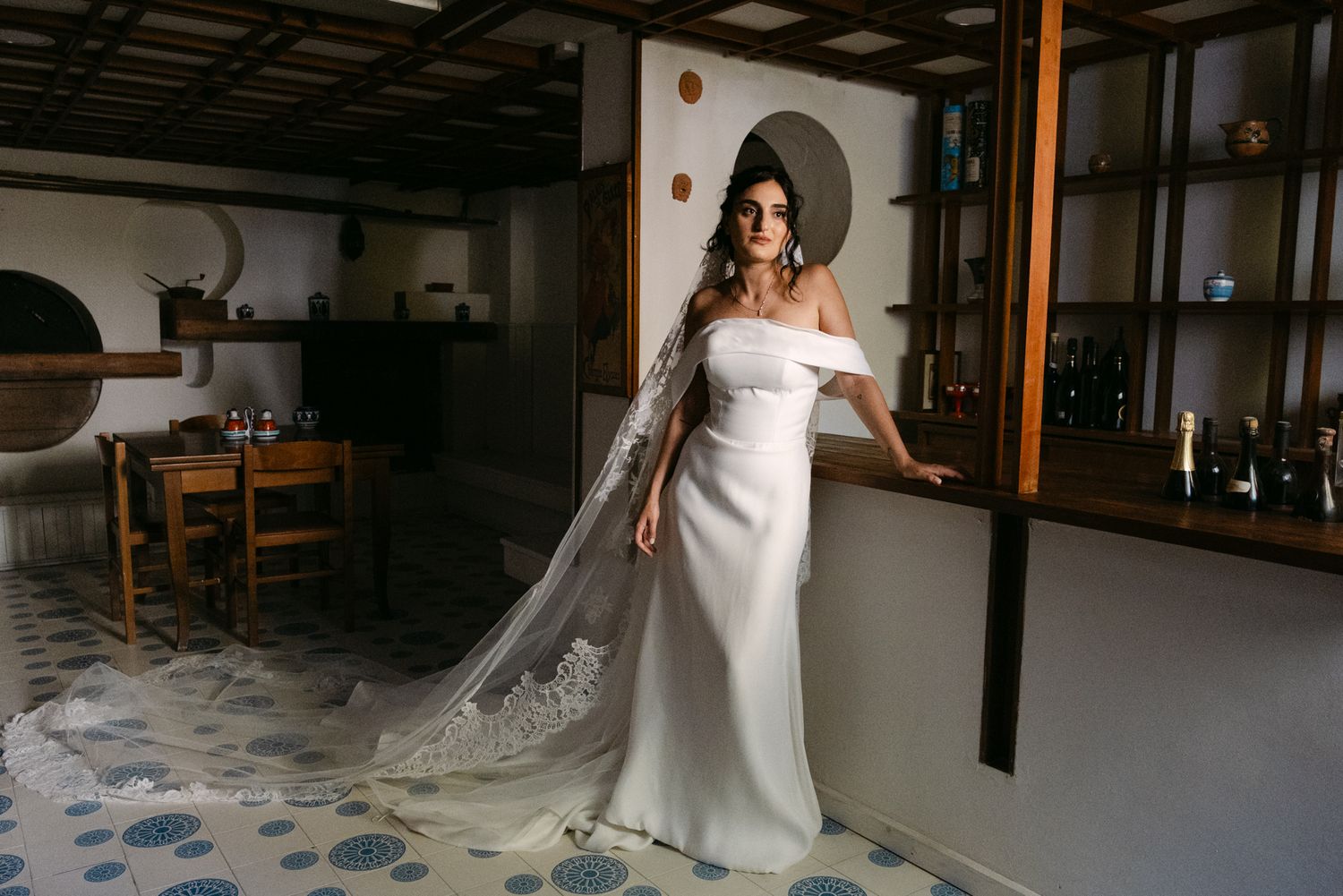 Bride in form-fitting off-shoulder gown with cathedral veil in rustic interior.