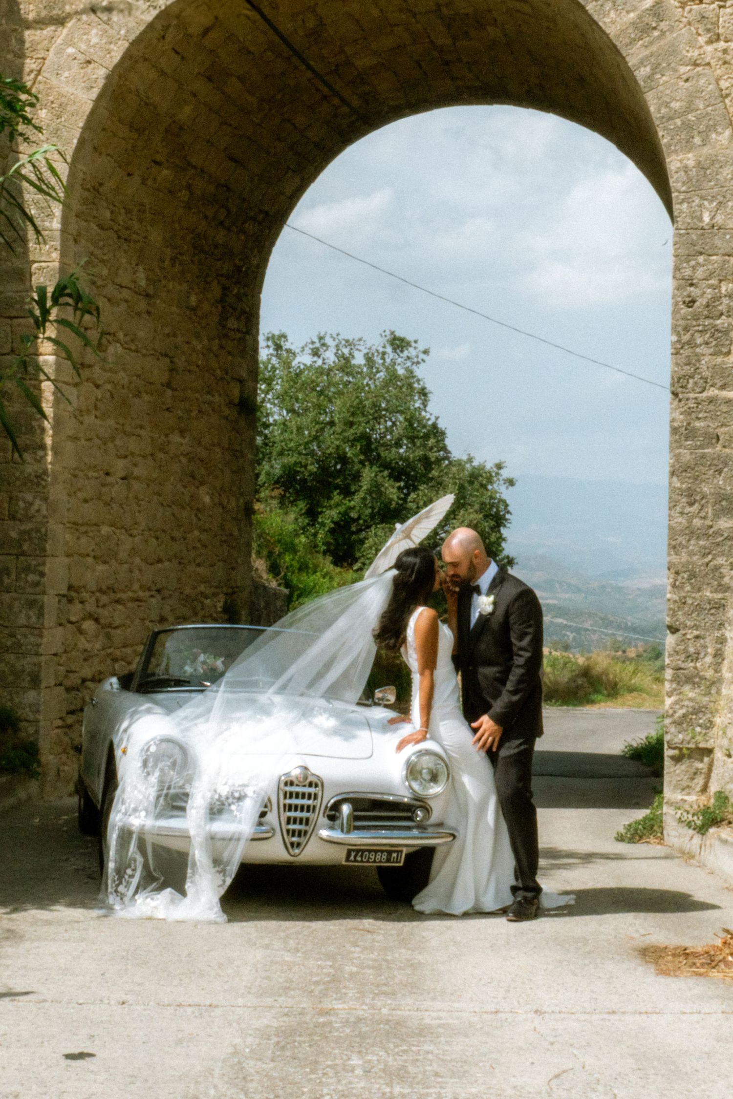 A romantic wedding photo sequence showing a couple and vintage car under stone arches in a scenic Italian setting.