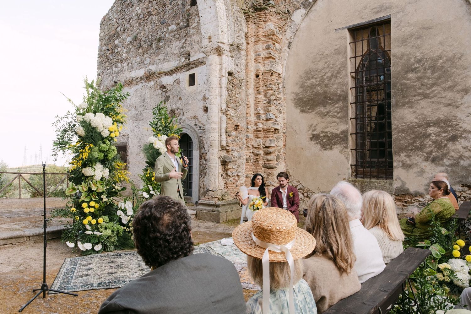 Guests mingle in an ancient stone venue decorated with lush greenery and white floral arrangements for a wedding.