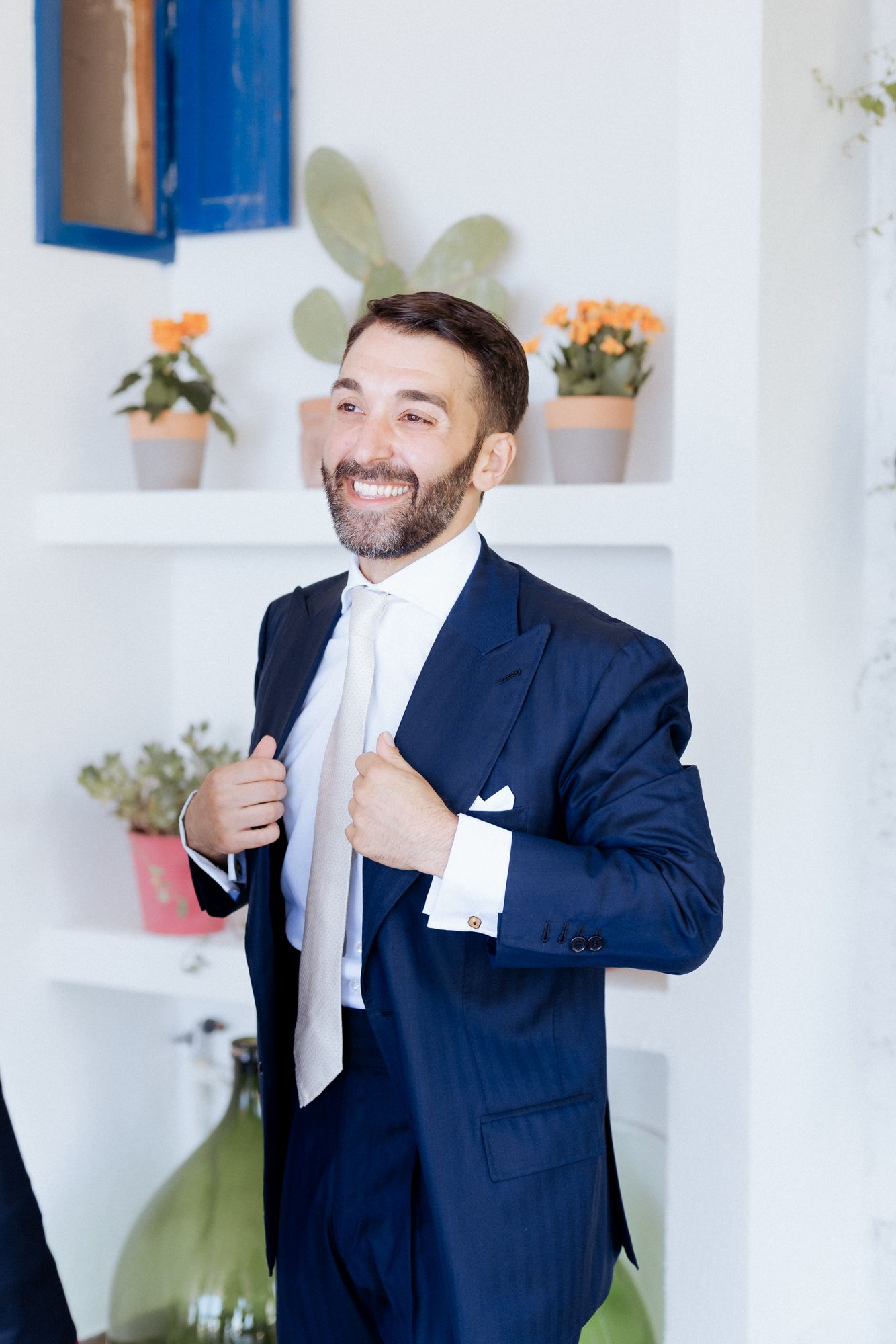 Groom in navy suit and white tie smiling in bright room with potted flowers.