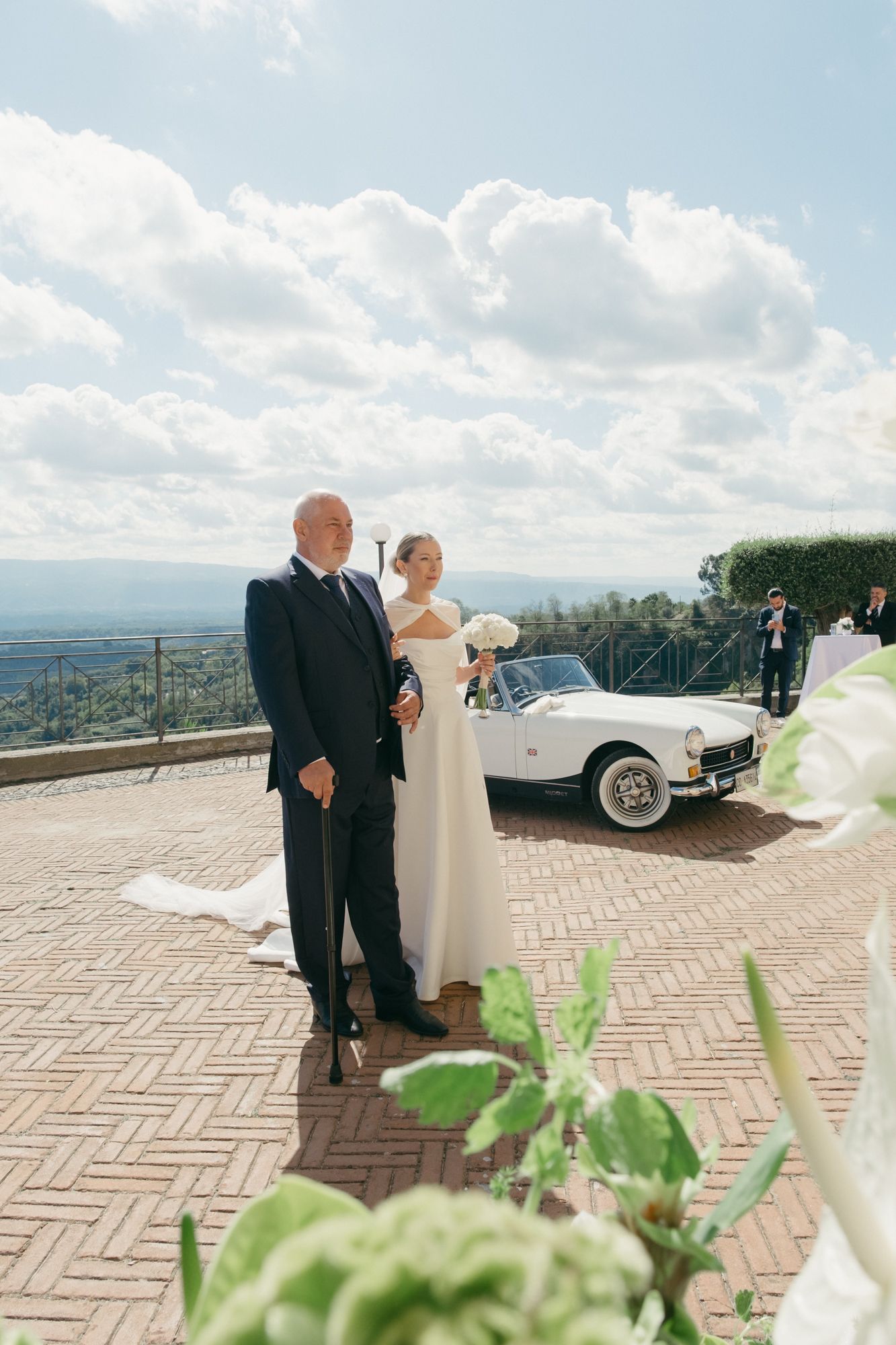 A wedding couple poses on a scenic brick terrace overlooking a panoramic view with a classic car in the background.