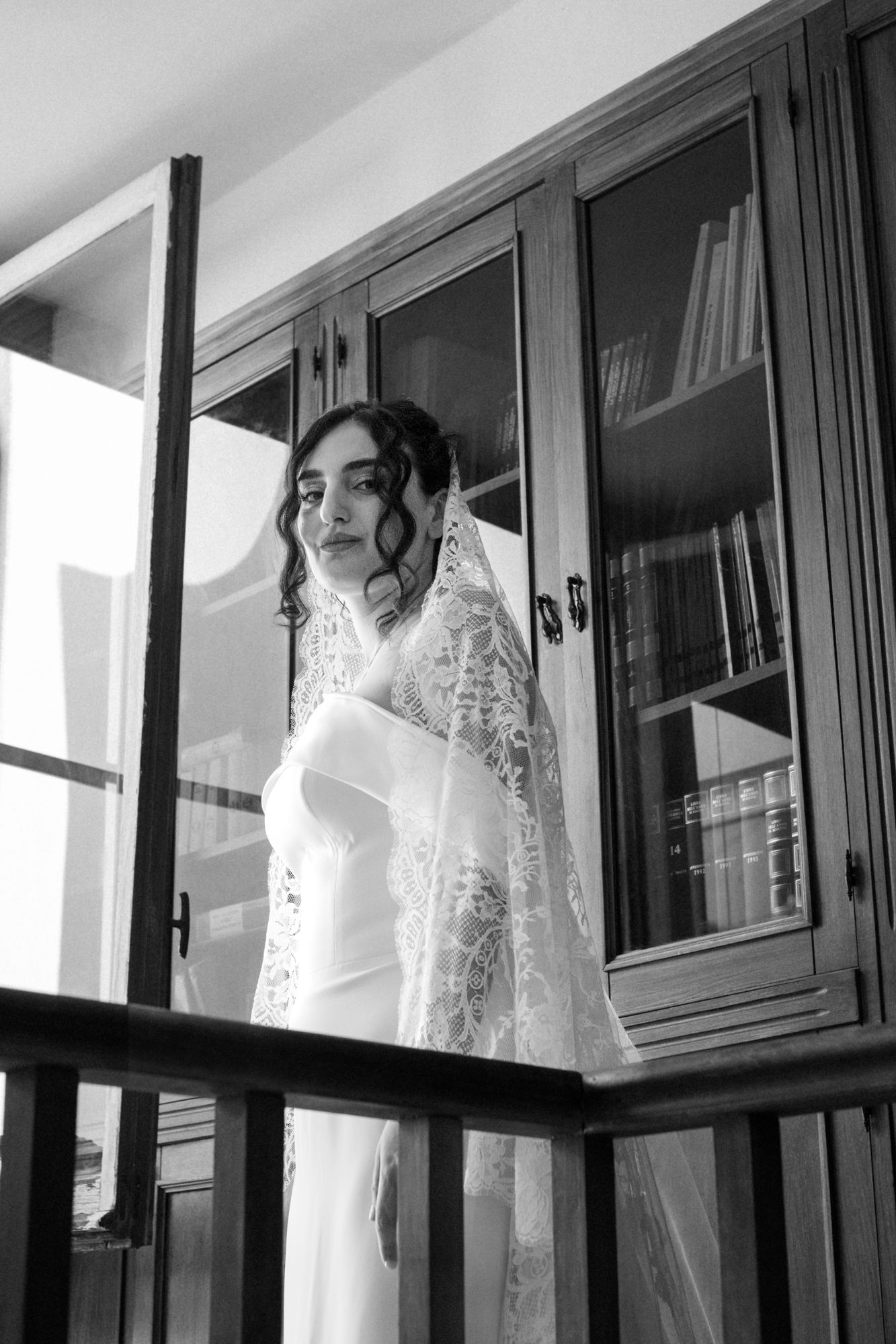 Black and white photo of bride in elegant dress standing behind glass cabinet.