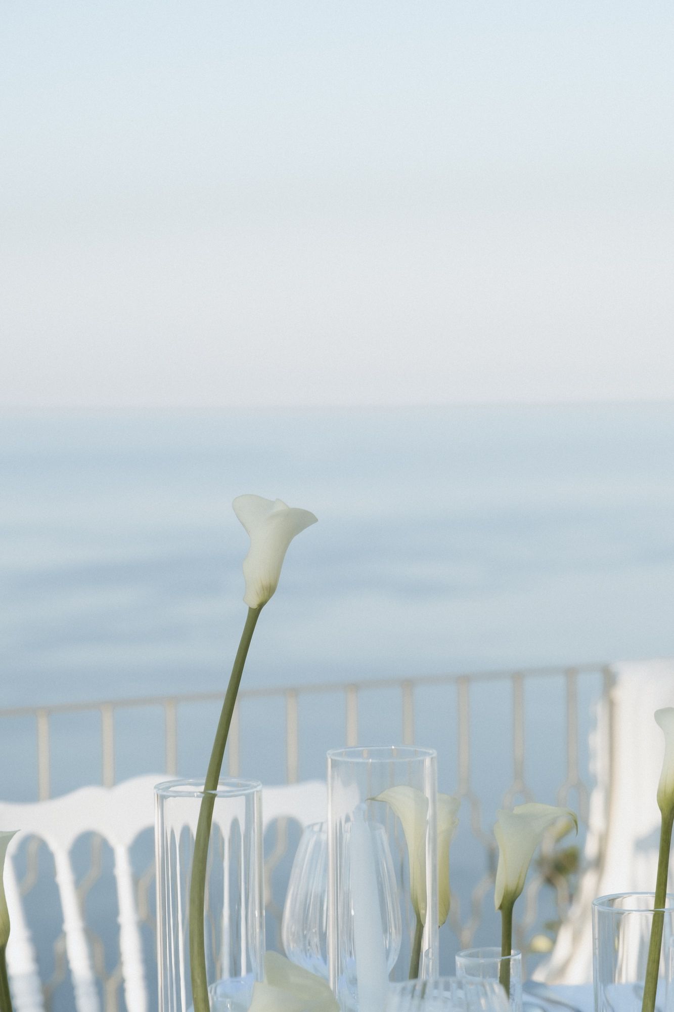 Row of empty wine glasses and white calla lilies set against a soft blue sky background.