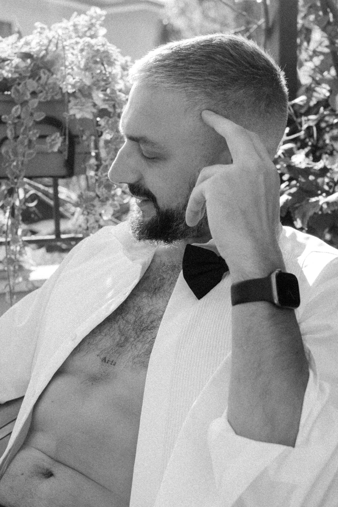 A bearded man in a white shirt and bowtie touches his temple while wearing a smartwatch.