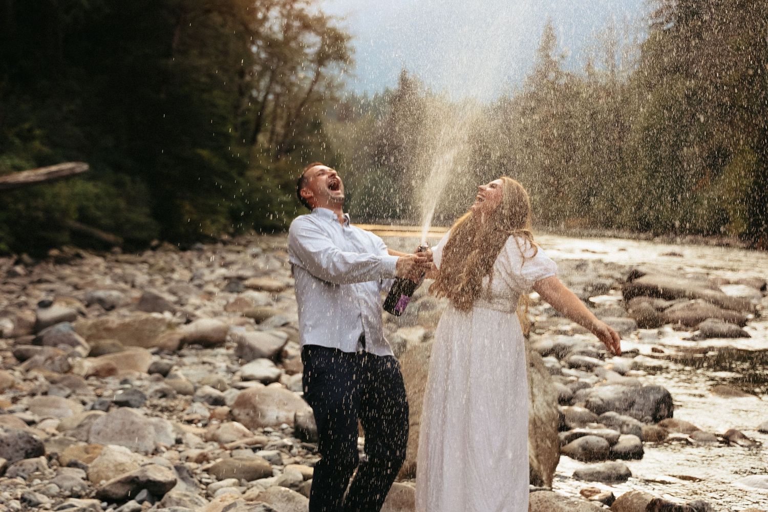 Playful couple splashes water at each other by rocky riverbed.
