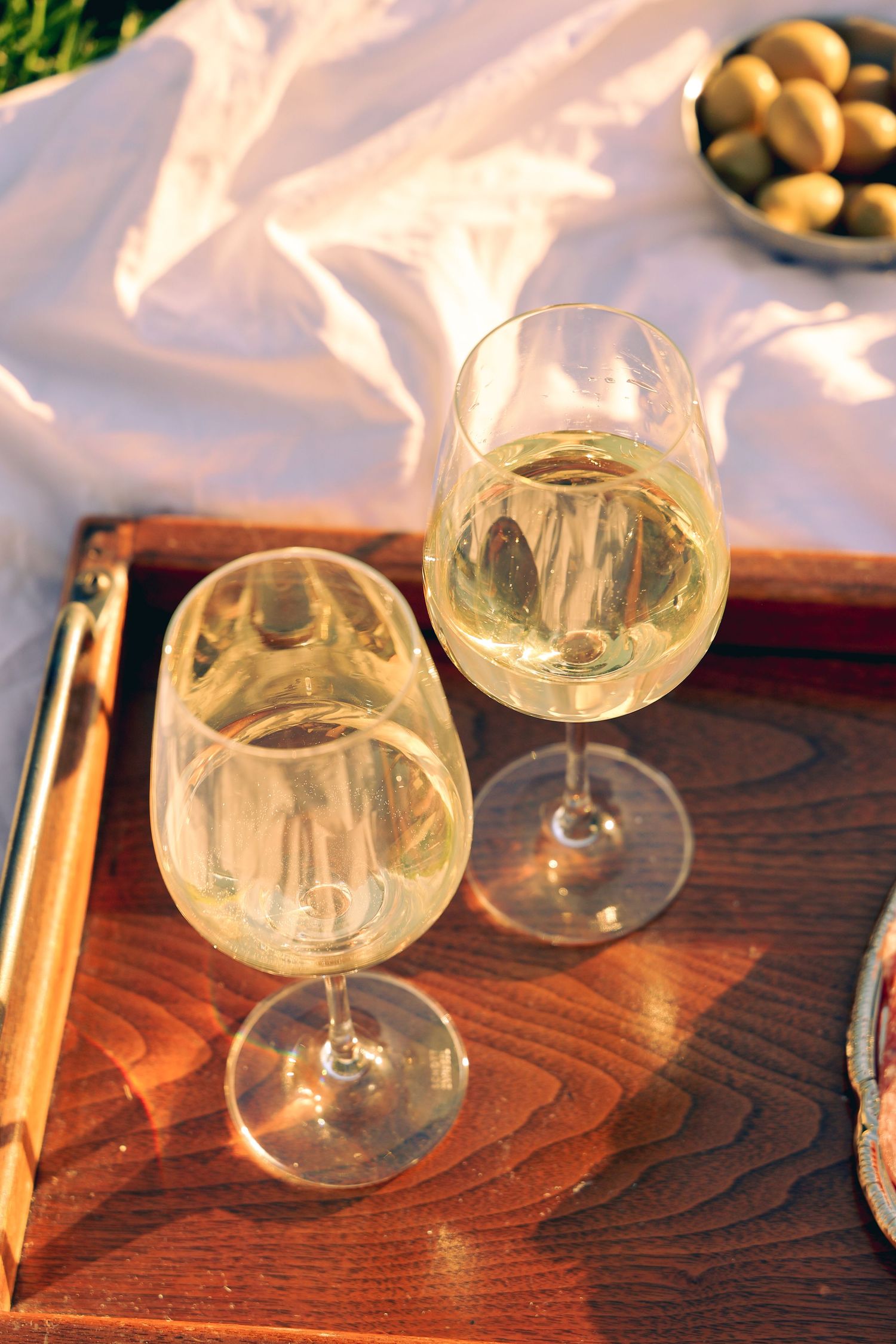 Two glasses of white wine on a wooden tray at a sunny outdoor picnic with olives and white cloth.