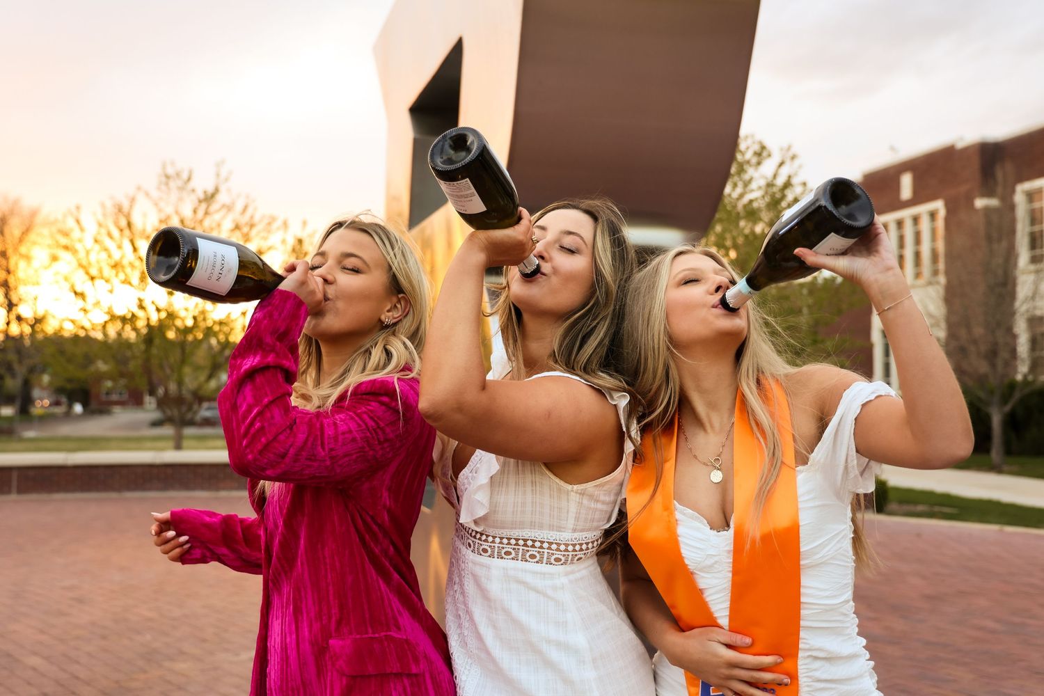 Group celebrating graduation by popping champagne bottles outdoors at sunset.