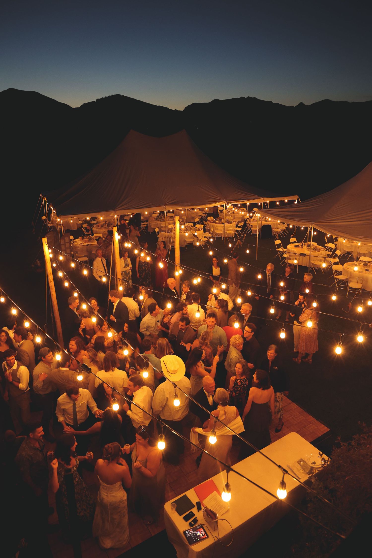 An evening wedding reception with glowing lanterns and guests gathered under mountain silhouettes.