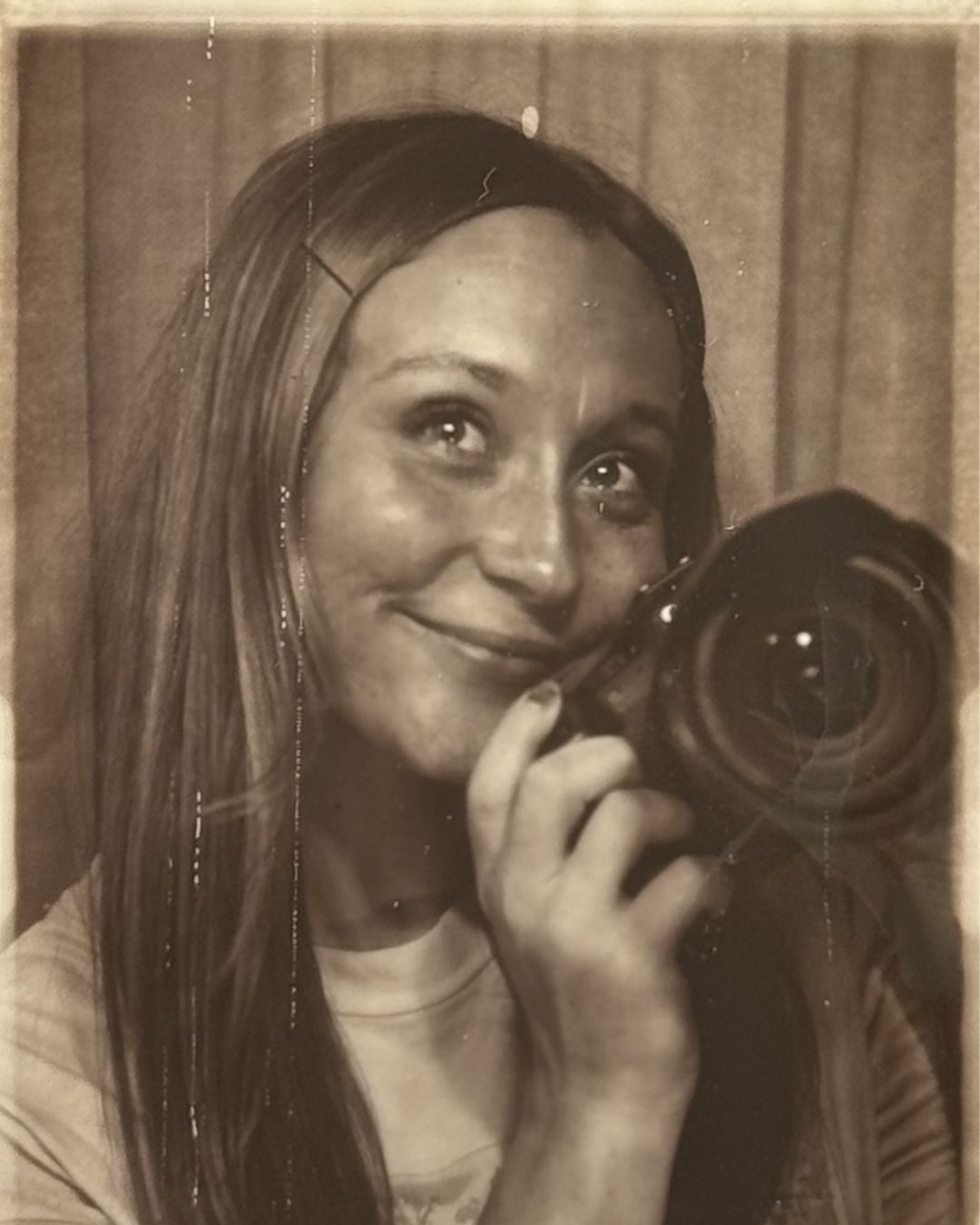 Vintage sepia photo of a smiling young woman holding a camera up to her face, long hair, retro photography aesthetic.