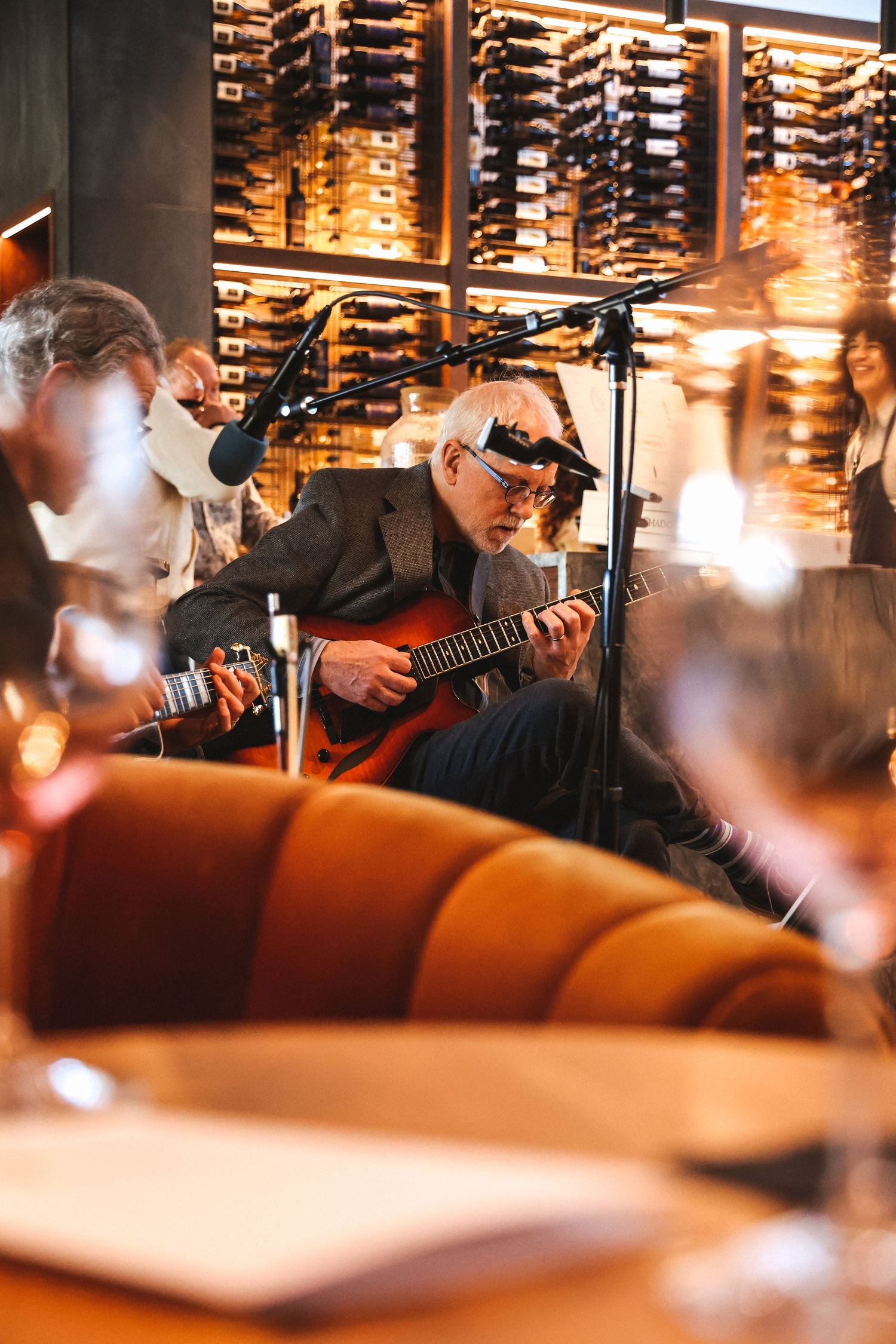 Musicians perform in a dimly lit venue with warm lighting and wine bottles displayed on shelves in the background.